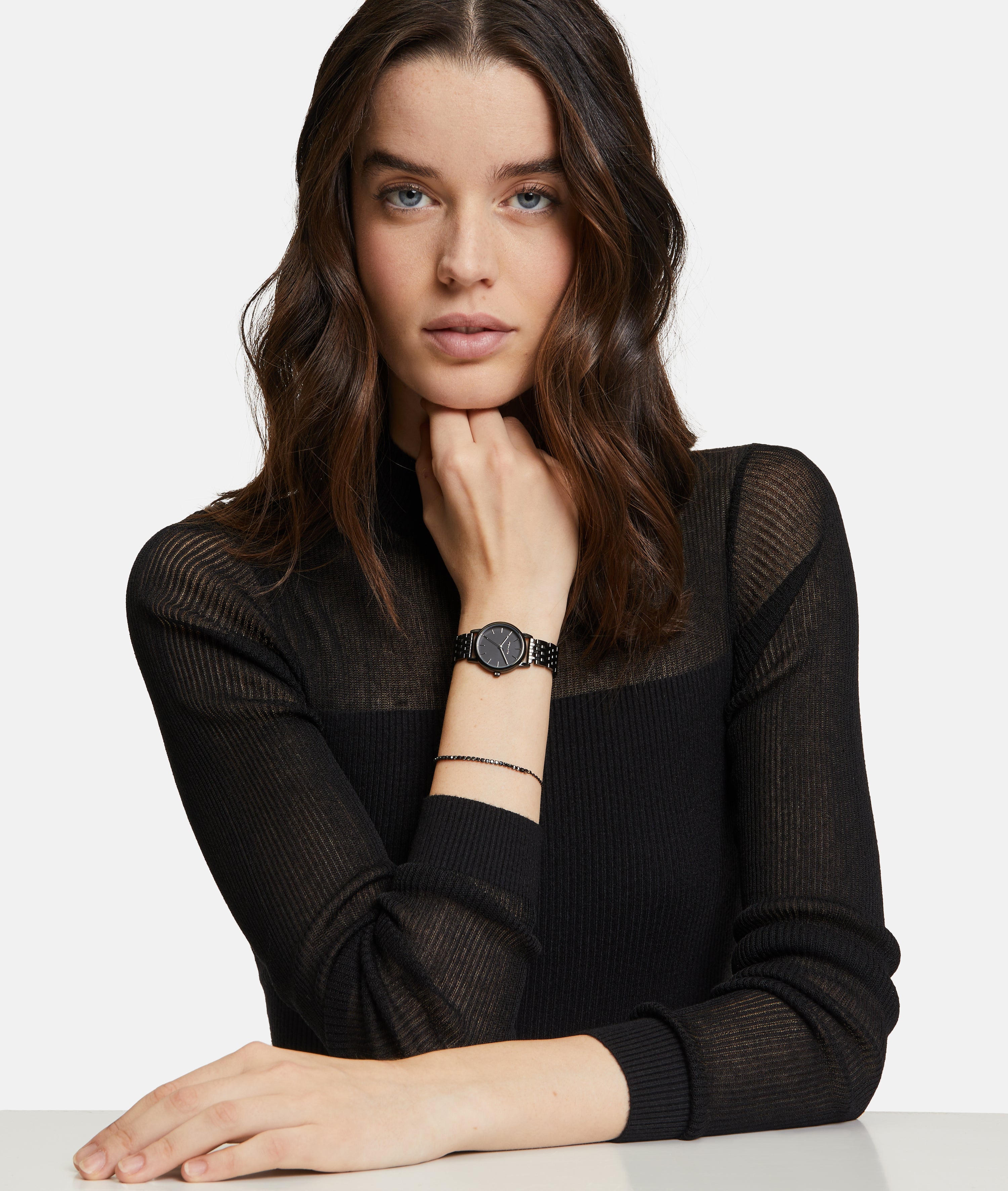 Eine Frau mit gewellten braunen Haaren in einem schwarzen Top lehnt sich an einen Tisch und betont das Tamaris Armband - The Black Sparkling Beads von Tamaris für einen schicken Lagenlook.