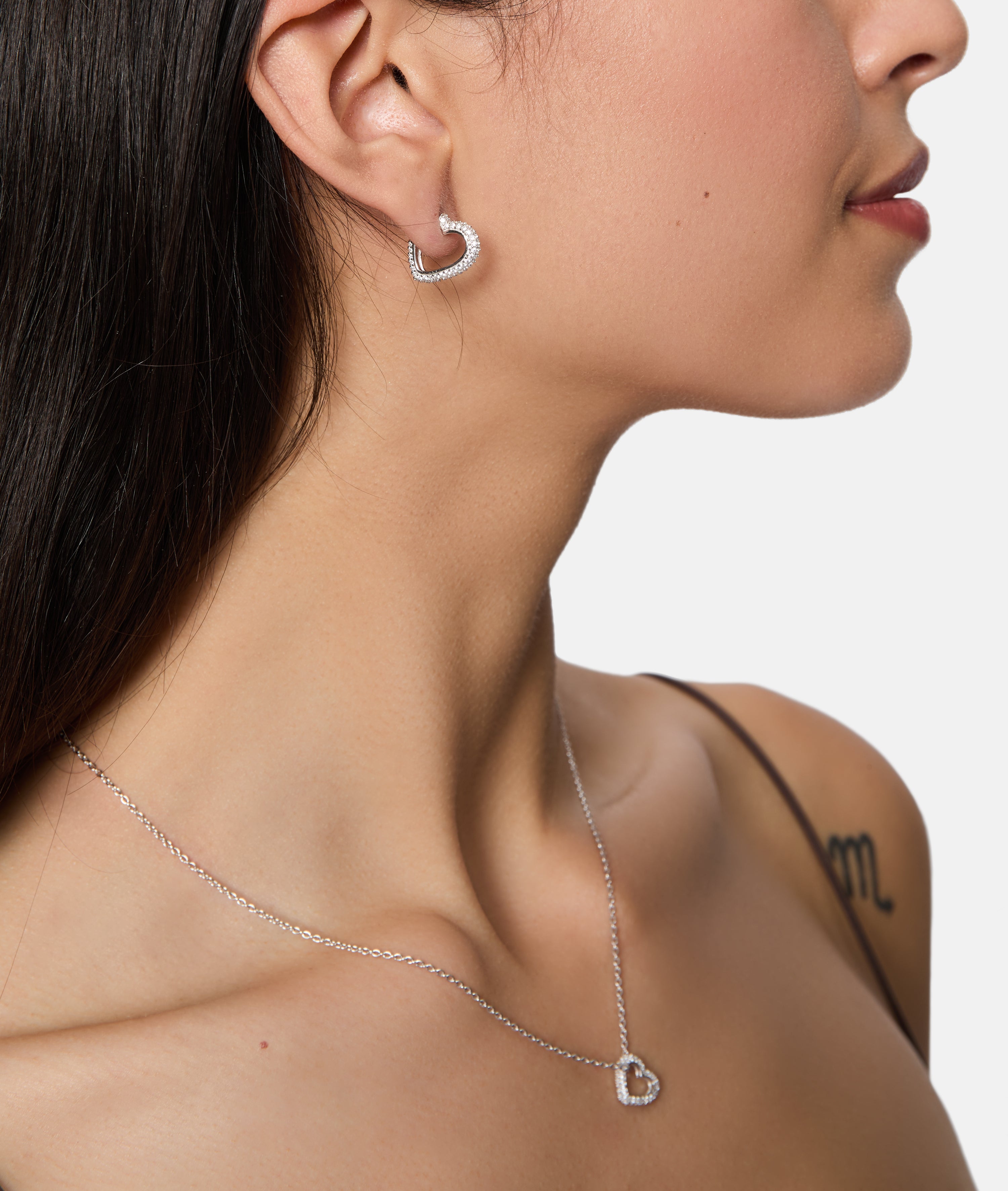 Eine Frau trägt Tamaris Creole - Sparkling Hearts 925 Silber Herz-Ohrringe mit Zirkonia und eine passende Herz-Anhänger-Halskette von Tamaris, ihr Schulter-Tattoo ist sichtbar.