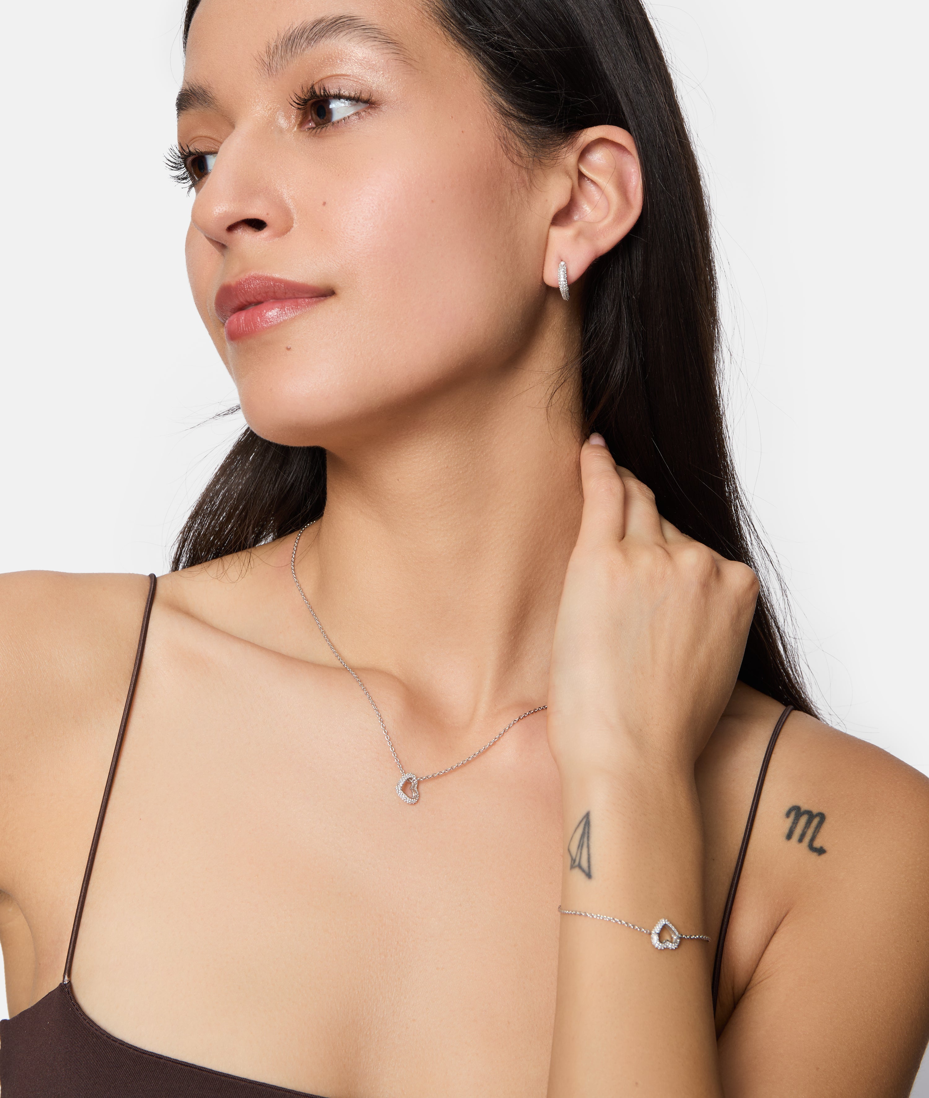 Eine Frau mit sichtbaren Tattoos trägt silberne Creolen, eine mit verspielten Zirkoniasteinen besetzte Herzanhänger-Halskette und das Tamaris Armband - Sparkling Hearts von Tamaris.