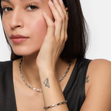 Eine Frau mit langen dunklen Haaren, die die Tamaris Halskette - The Edge von Tamaris und eleganten Schmuck trägt, berührt ihr Gesicht und enthüllt ein Papierflugzeug-Tattoo auf ihrem Handgelenk.