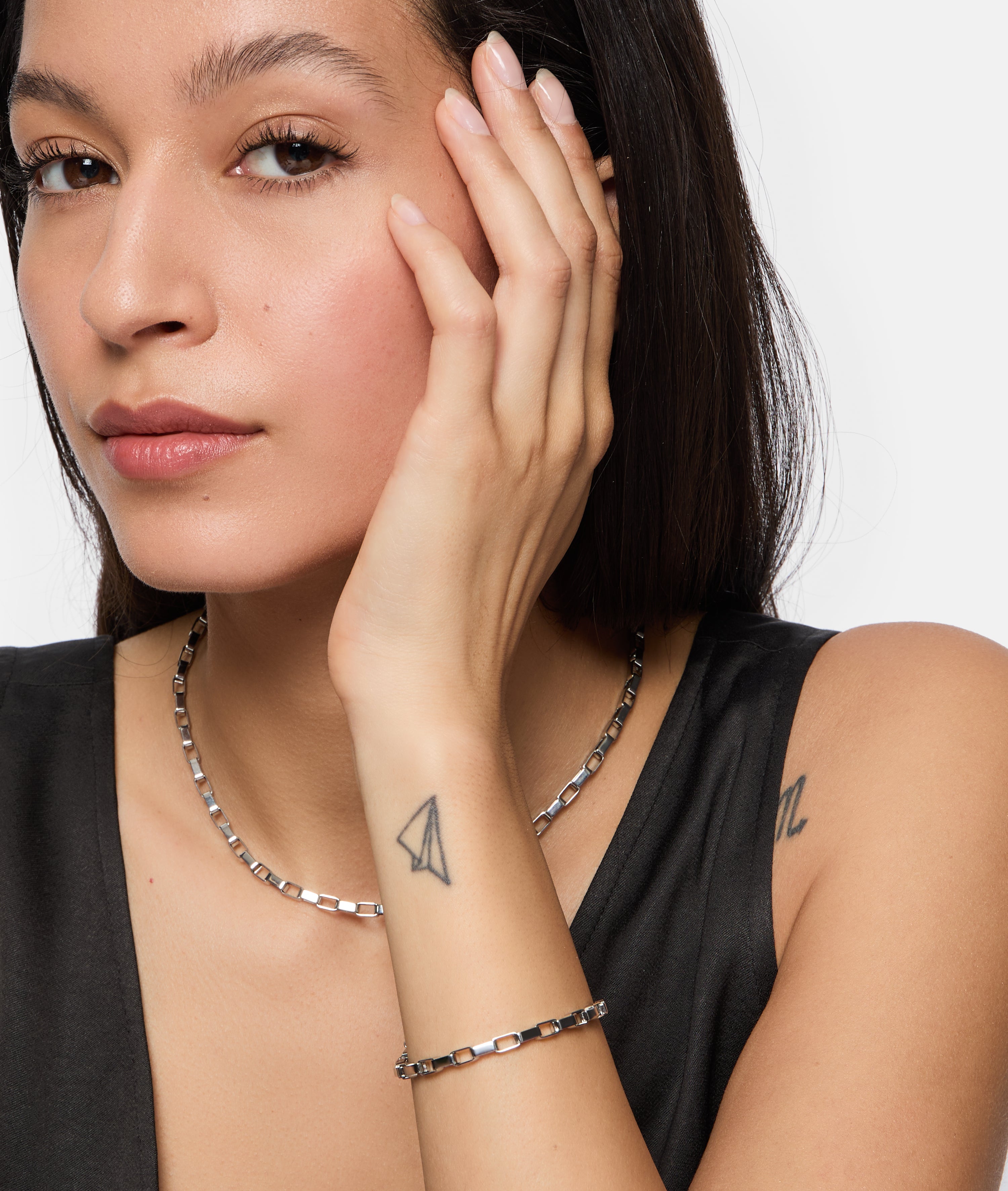 Eine Frau mit langen dunklen Haaren, die die Tamaris Halskette - The Edge von Tamaris und eleganten Schmuck trägt, berührt ihr Gesicht und enthüllt ein Papierflugzeug-Tattoo auf ihrem Handgelenk.