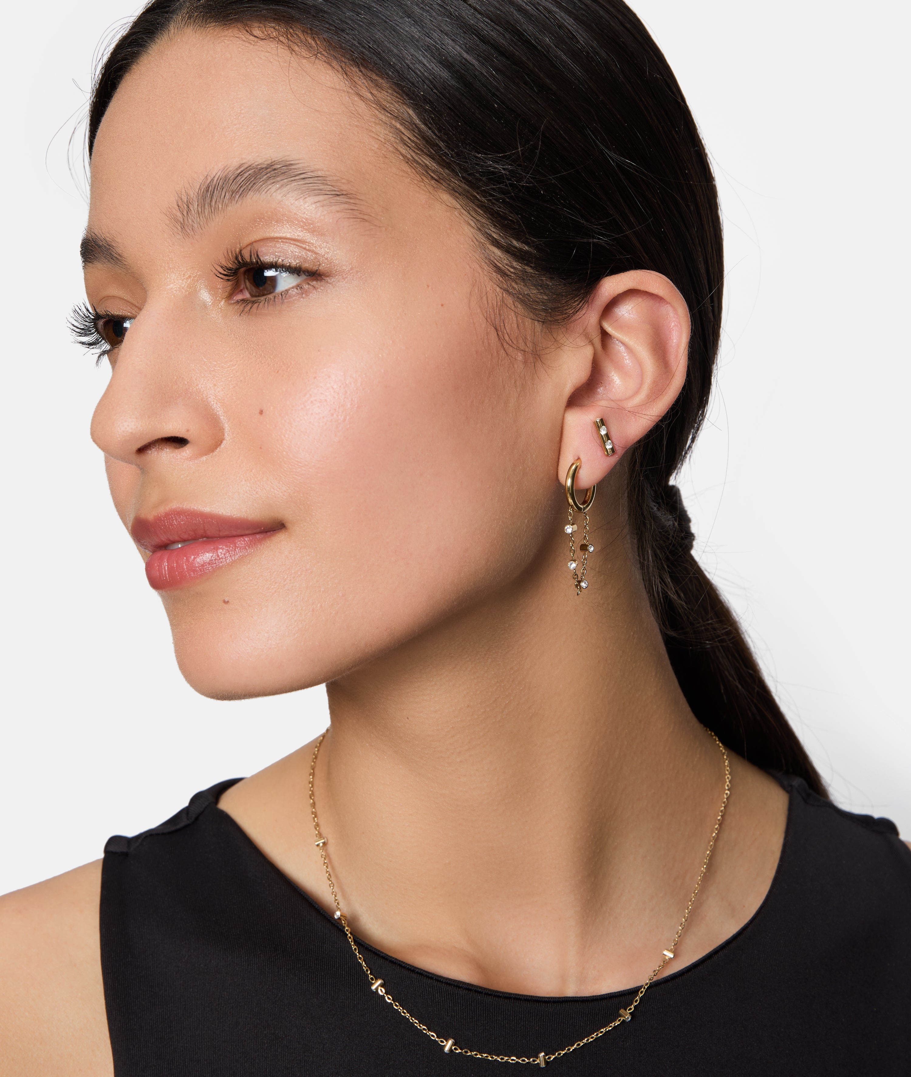 Frau mit langen dunklen Haaren trägt Tamaris Ohrstecker - Tiny Tubes und eine goldene Halskette, bekleidet mit einem ärmellosen schwarzen Top - ihr Tamaris-Schmuck glänzt mit eleganten Details.