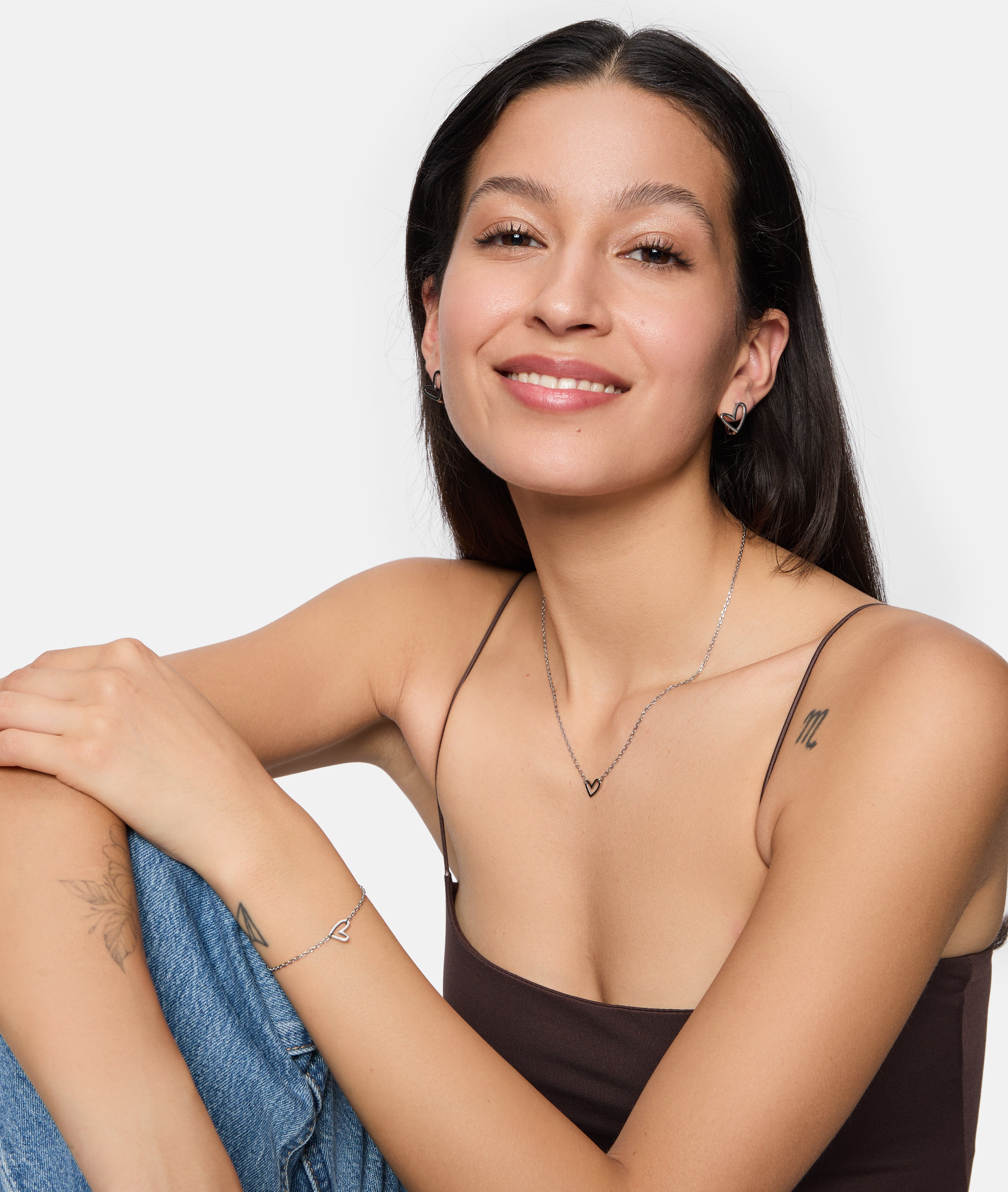 Lächelnde Frau mit langen dunklen Haaren trägt Tamaris Creole - Graffiti Heart Ohrringe, ein braunes Top und Blue Jeans, während sie lässig sitzt.
