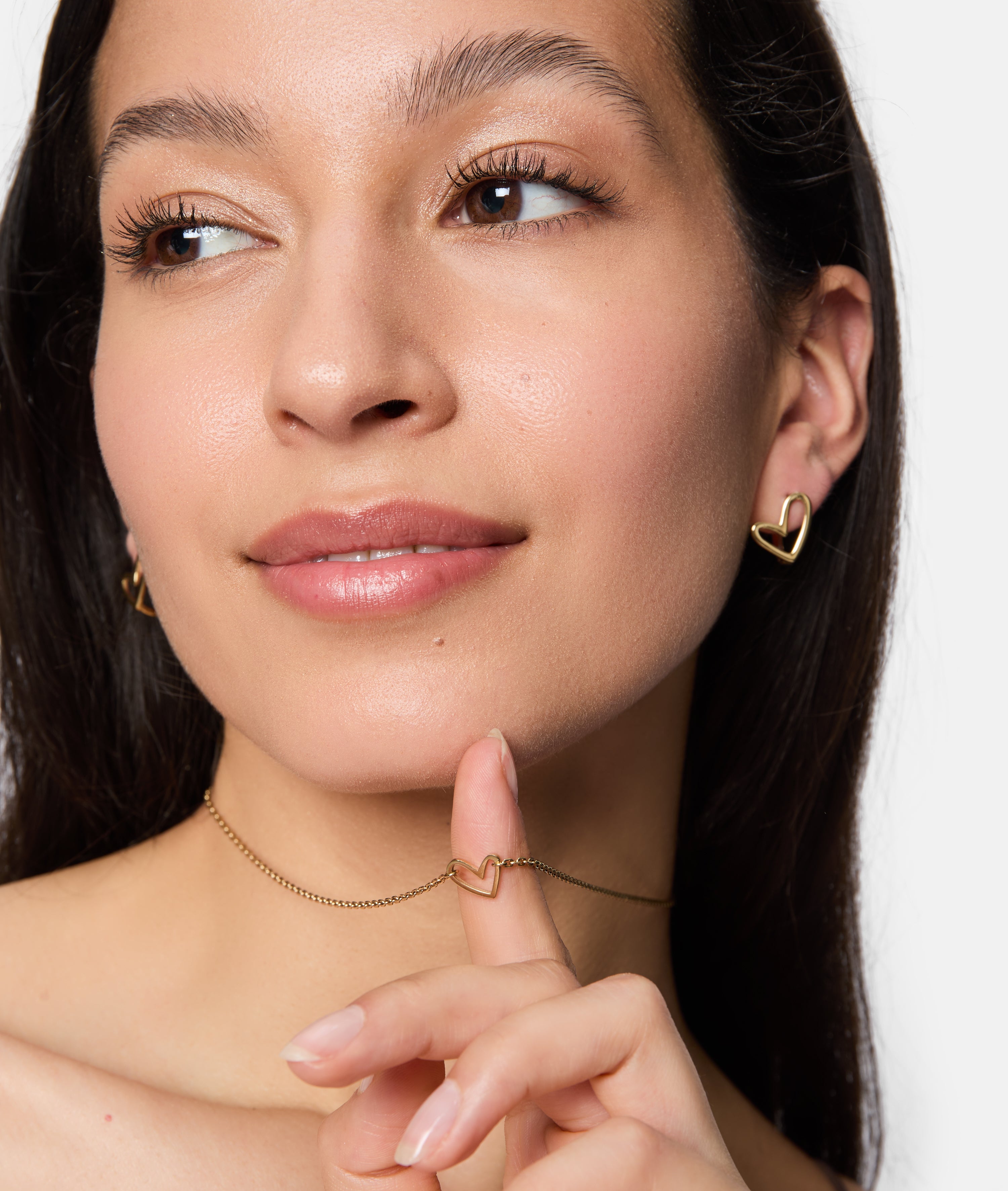 Eine Frau lächelt, trägt goldene Ohrringe in Herzform und die Tamaris Halskette - Graffiti Heart, die sanft die Kette berührt - dieses auffällige Stück von Tamaris ist das perfekte Accessoire für Frauen.