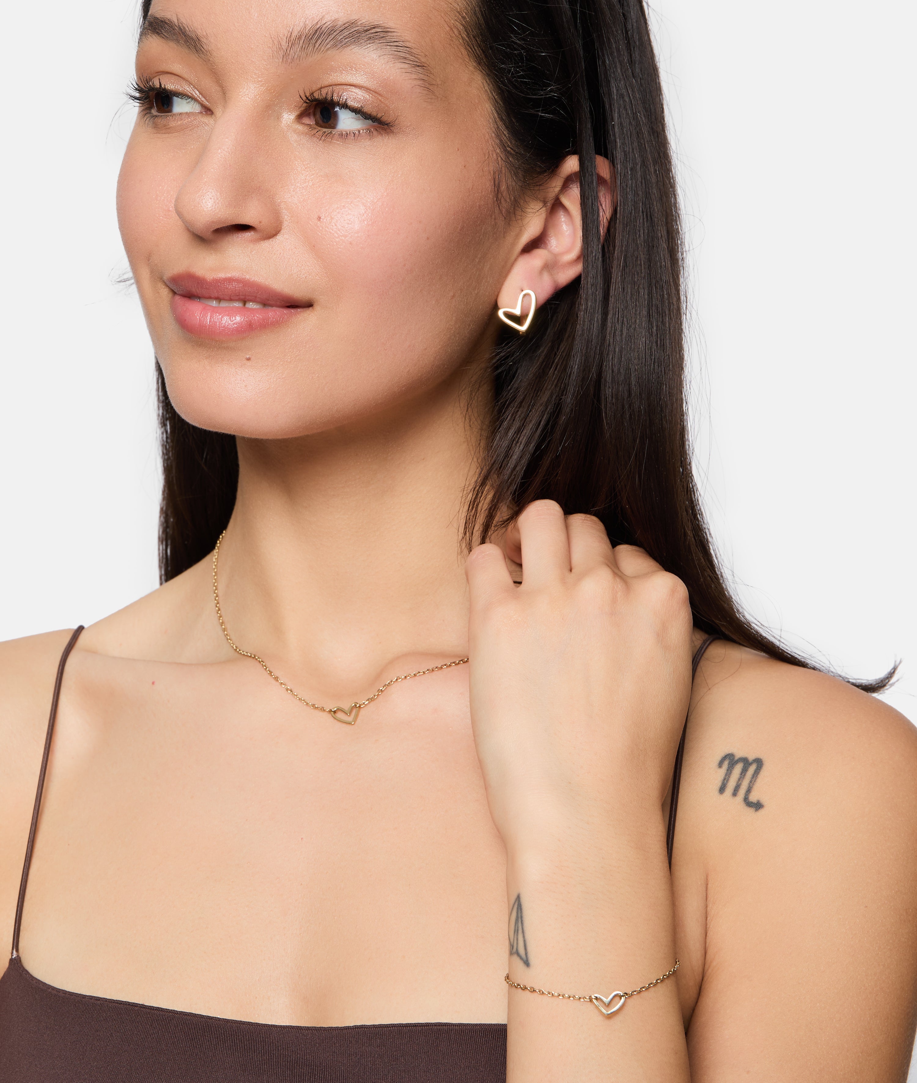 Eine Frau mit Tattoos auf Arm und Schulter trägt goldene Ohrringe in Herzform, ein Armband und die Tamaris Halskette - Graffiti Heart, die ihrem Accessoire Frauen-Look einen trendigen Touch verleiht.