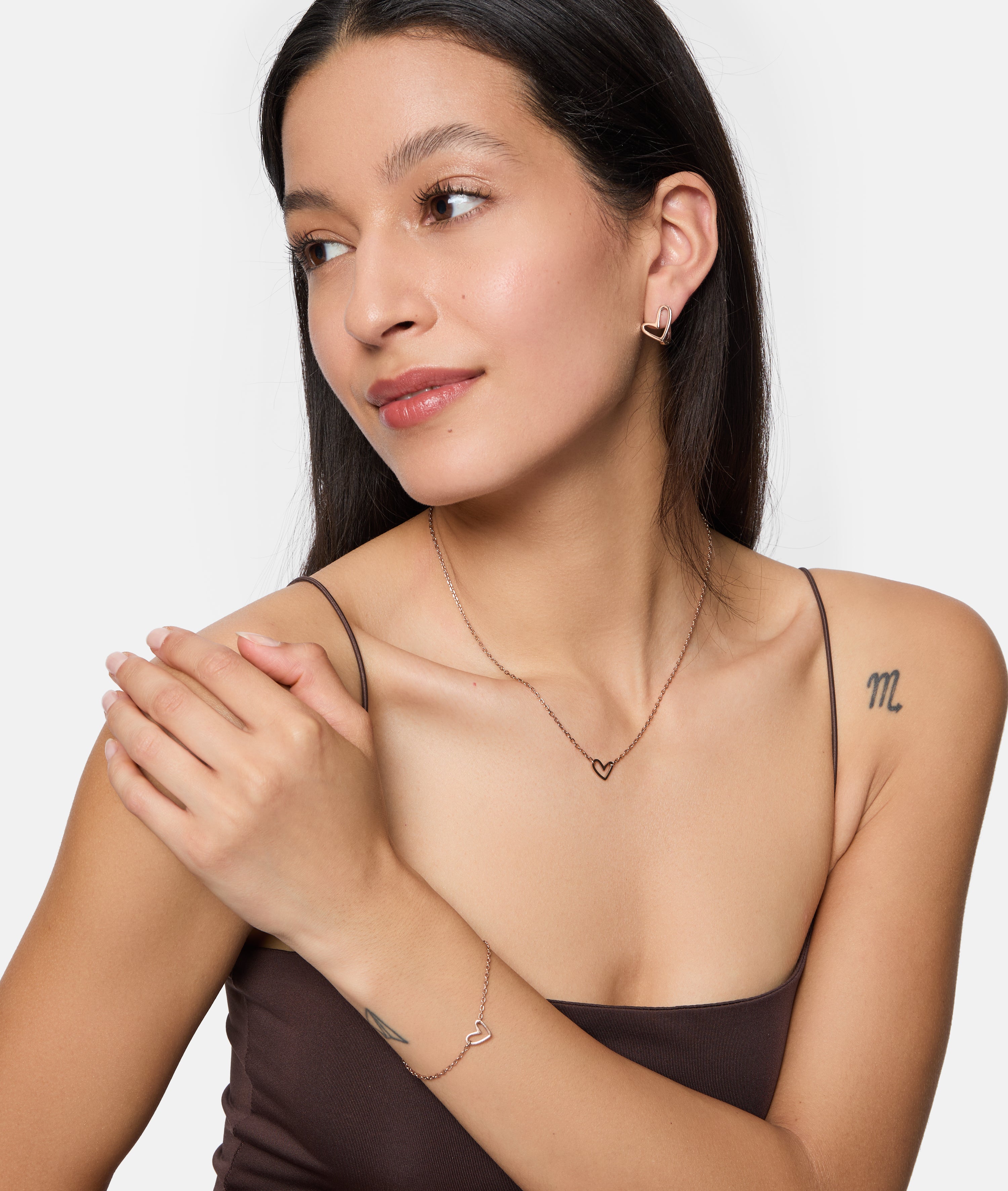 Frau trägt die Tamaris Creole - Graffiti Heart Ohrringe, Teil eines herzförmigen Schmucksets von Tamaris, bestehend aus Halskette und Armband, gepaart mit einem braunen Top und sichtbaren Tattoos.