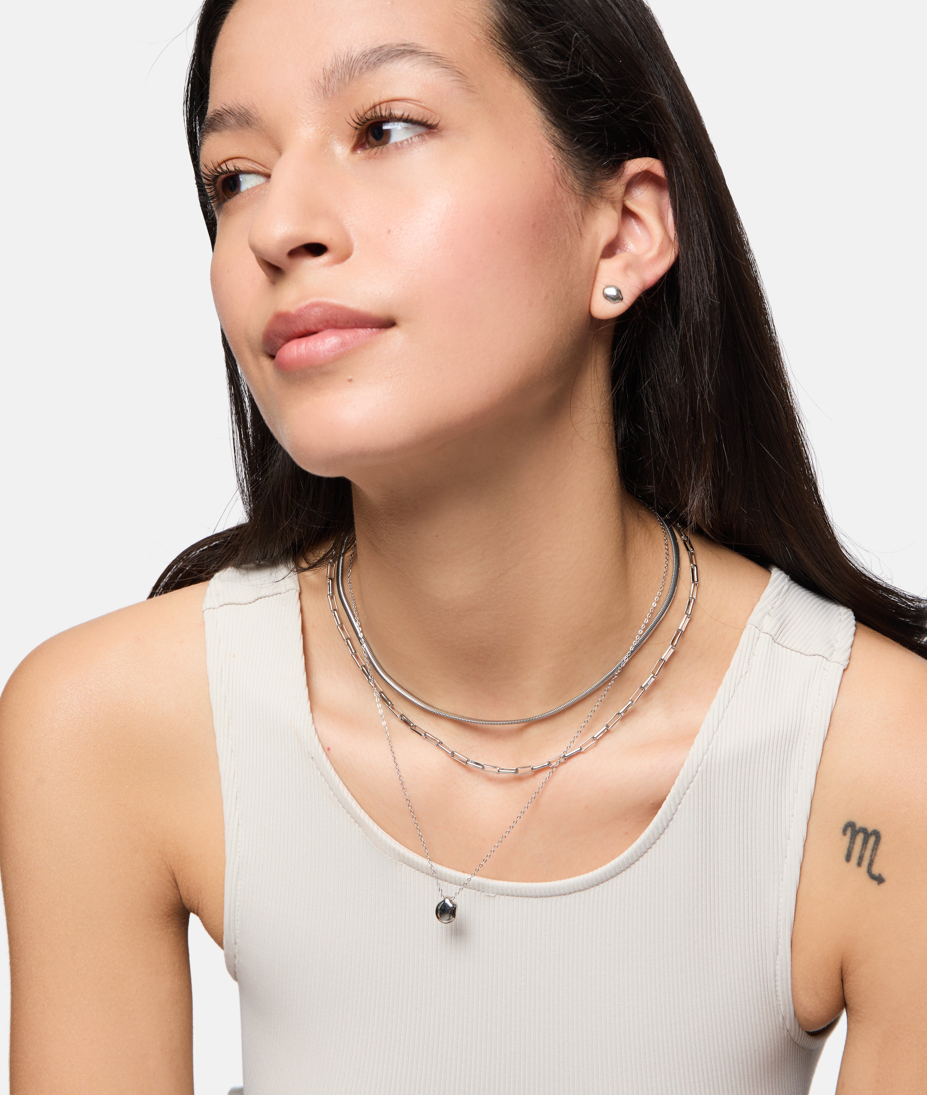Frau in weißem Tank-Top mit mehrlagigen Silberketten und Tamaris Ohrstecker - Modern Layering von Tamaris, mit organischem Design, sowie einem "m"-Tattoo auf dem Arm.