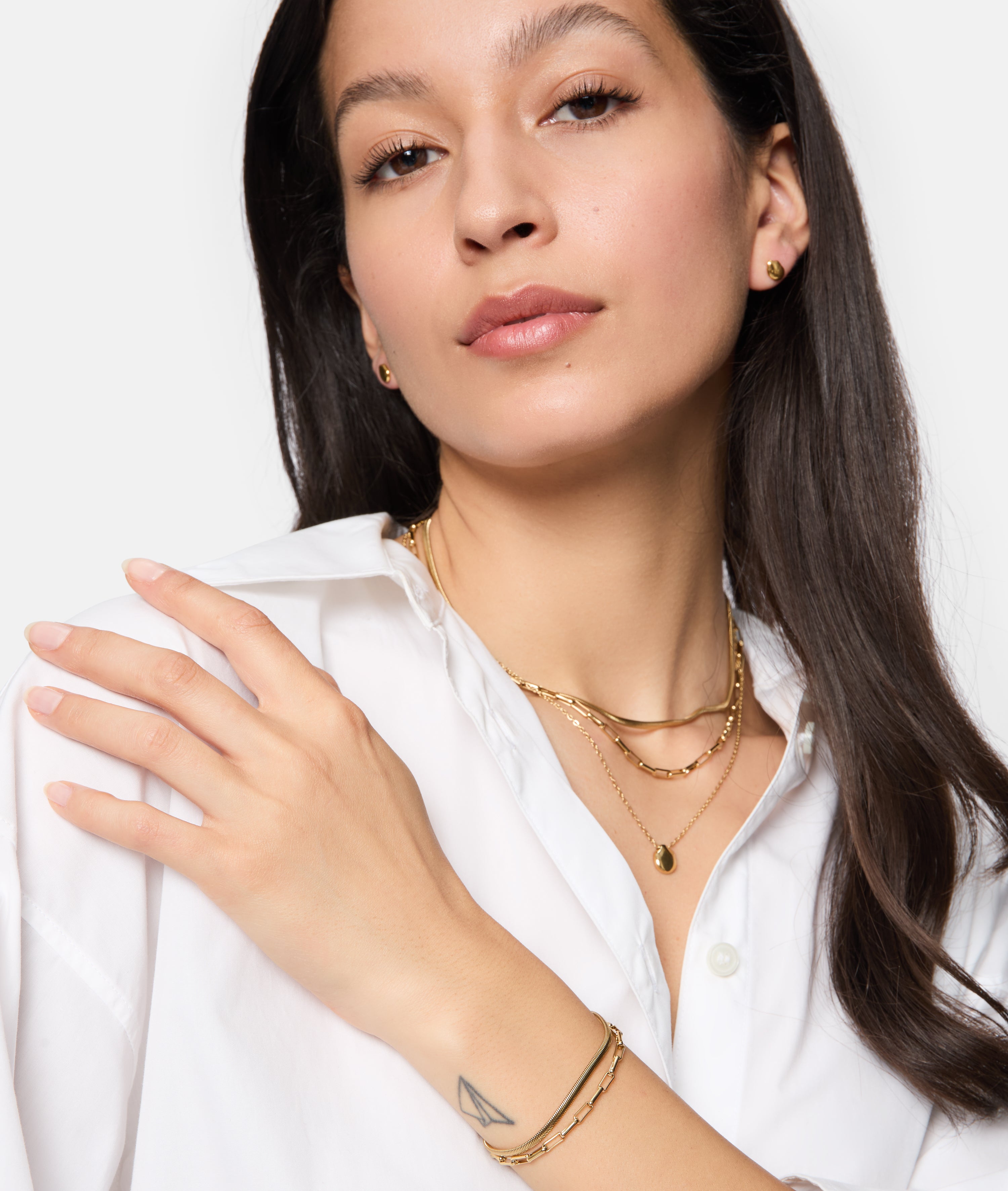 Eine Frau mit weißem Hemd trägt einen goldenen Tamaris Ohrstecker - Modern Layering mit organischem Muster, der sanft ihre Schulter berührt und ein kleines Dreieckstattoo an ihrem Handgelenk enthüllt, das ihren eleganten Look vervollständigt.