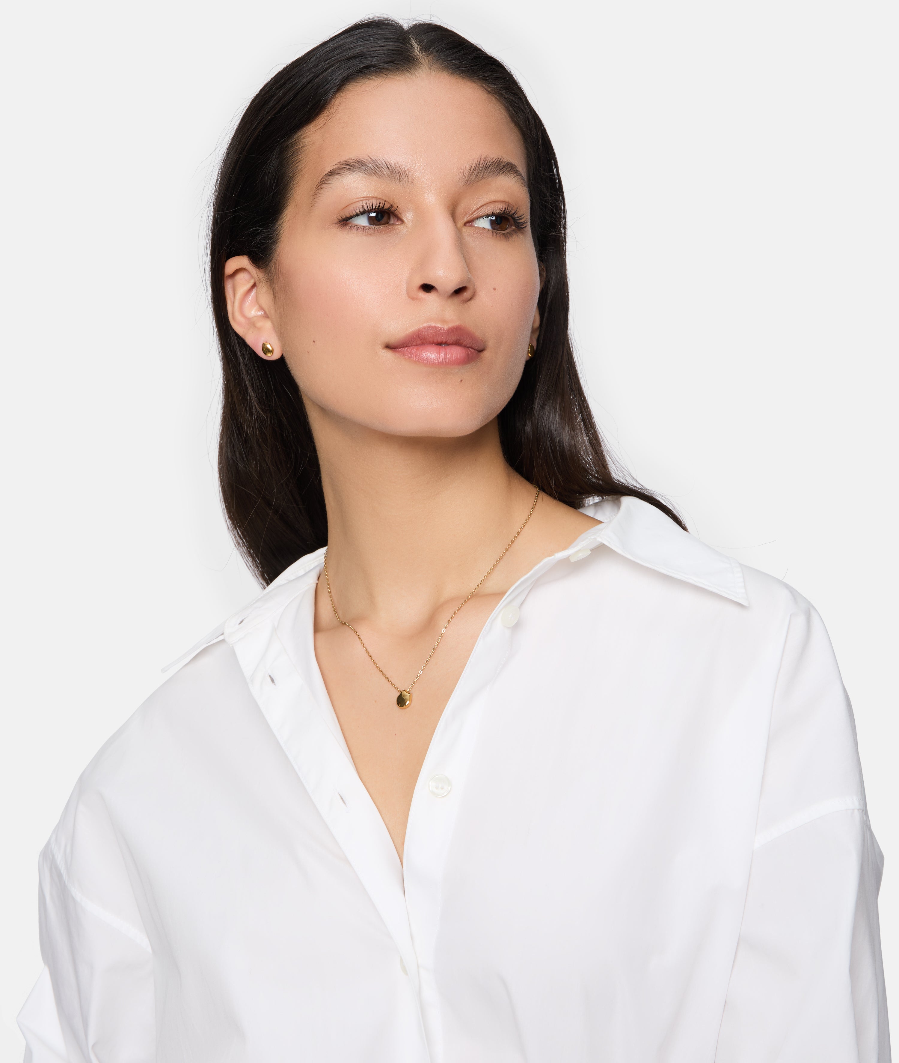 Frau im weißen Hemd trägt Tamaris Ohrstecker - Modern Layering by Tamaris und zwei goldene Halsketten mit organischem Muster, leicht zur Seite schauend.