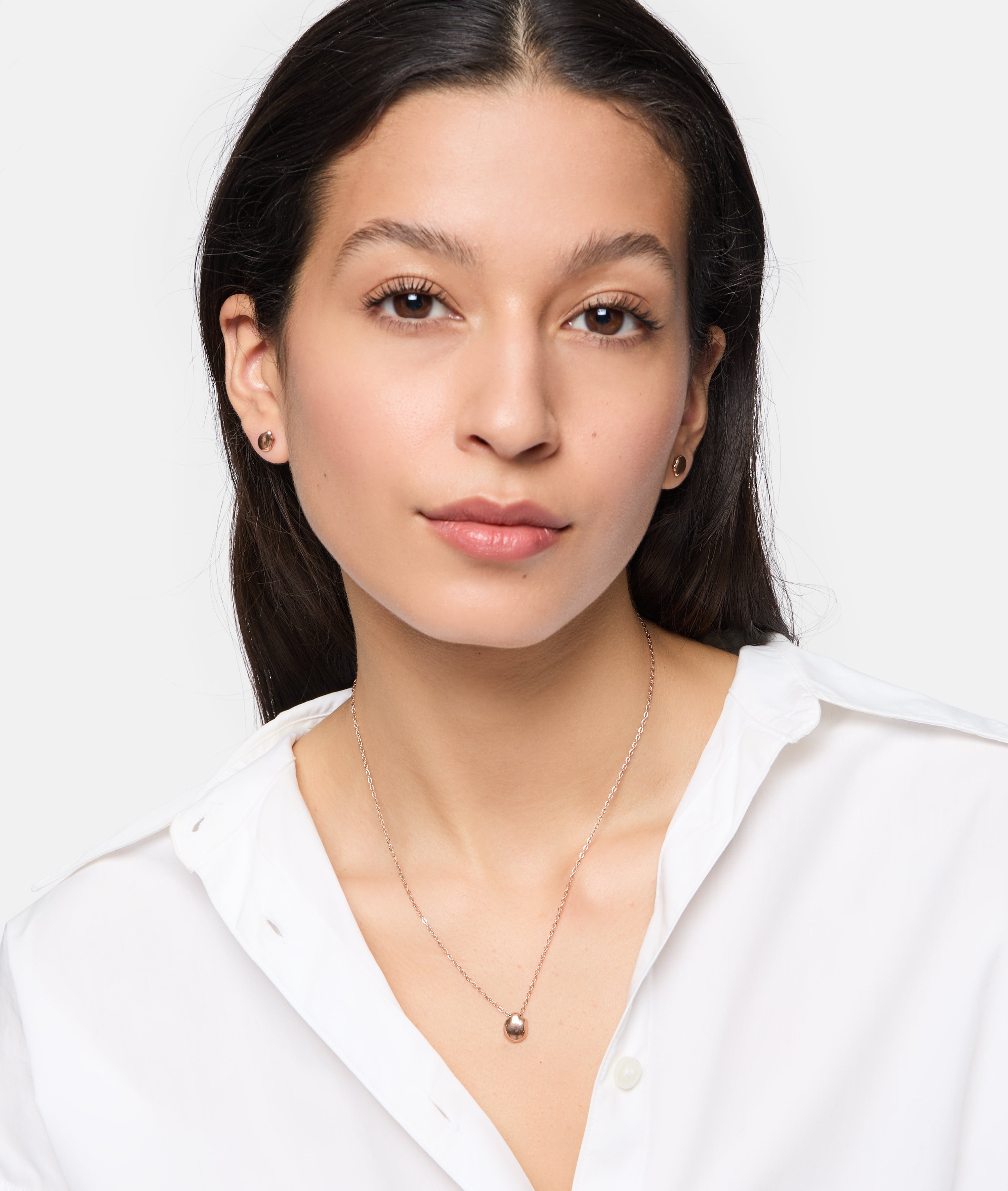 Frau mit langen dunklen Haaren und weißem Hemd trägt Tamaris Ohrstecker - Modern Layering by Tamaris und eine Anhänger-Halskette mit organischem Design und schaut in die Kamera.
