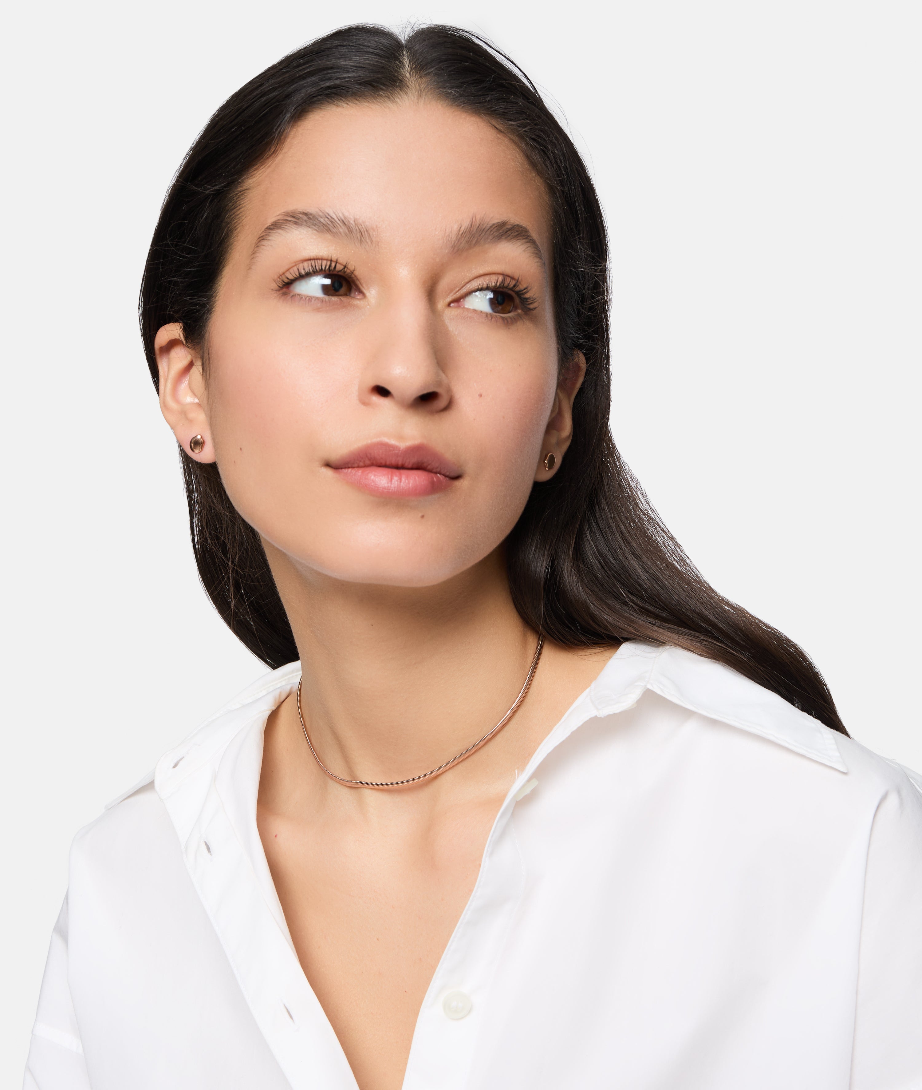 Frau mit langen dunklen Haaren trägt ein weißes Hemd, kleine Ohrstecker und die Tamaris Halskette - Modern Layering von Tamaris, mit Blick zur Seite.