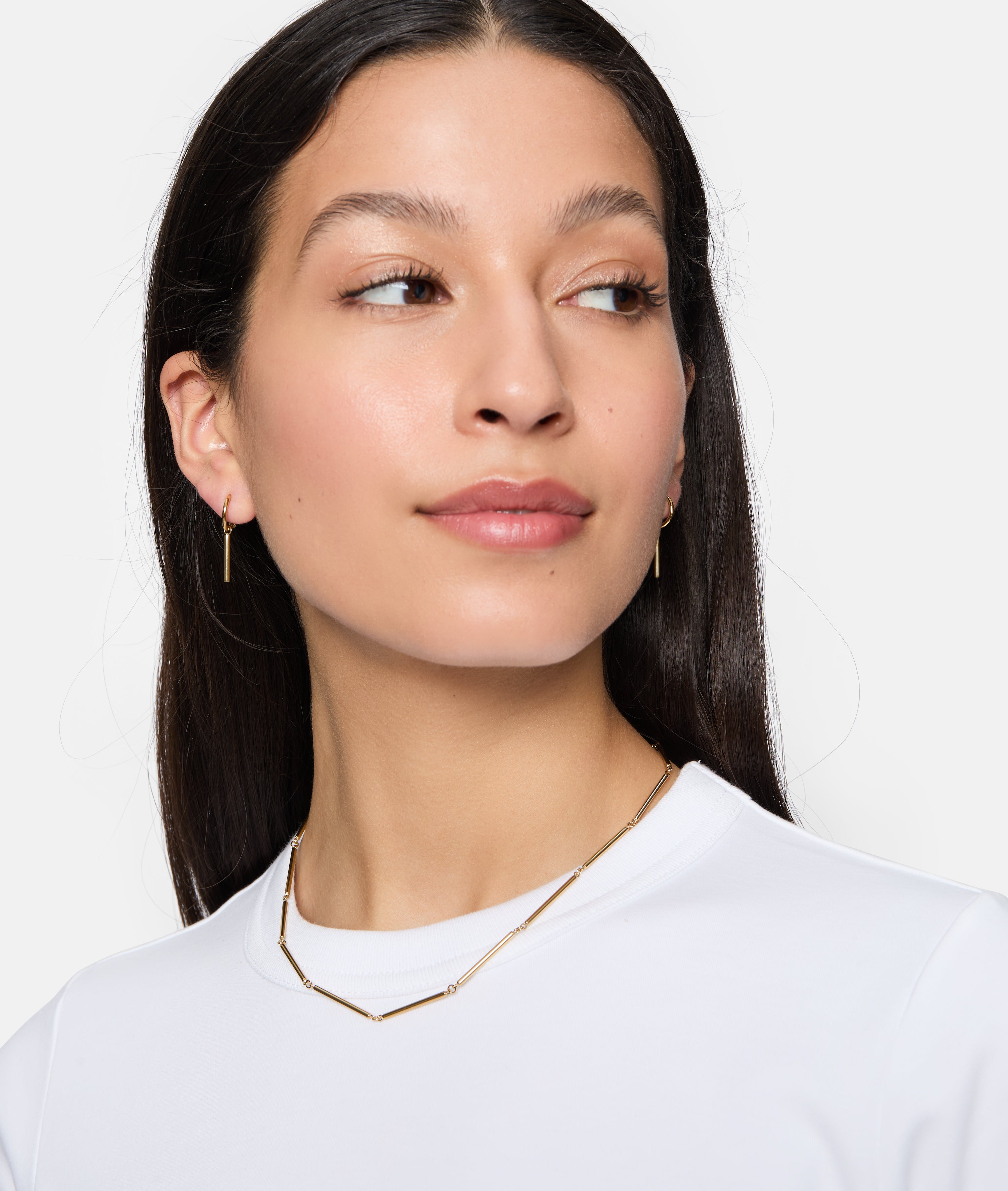 Eine Frau mit langen dunklen Haaren trägt die Tamaris Halskette - Modern Chain von Tamaris und ein weißes Oberteil, schaut leicht zur Seite und lächelt sanft.