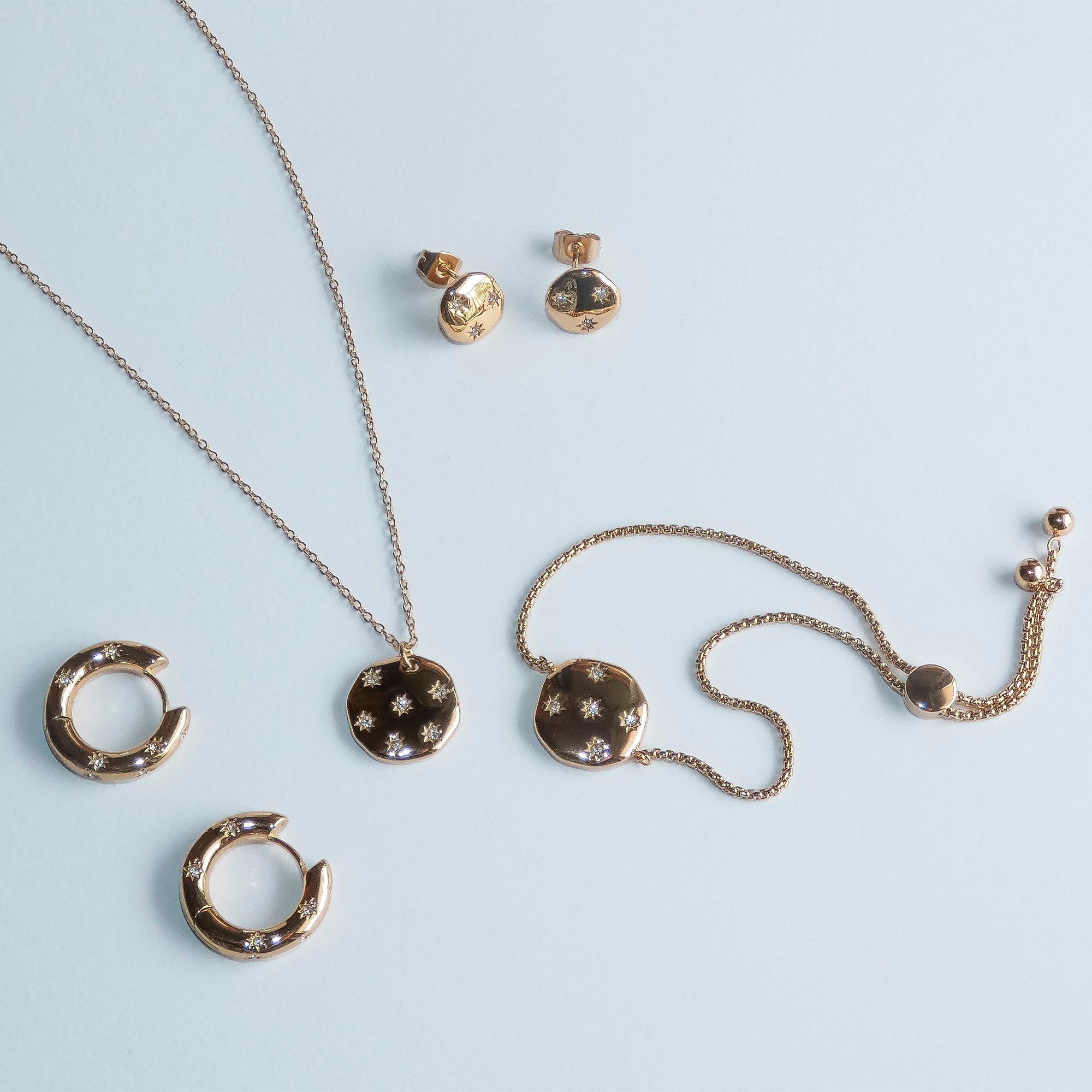 Die Tamaris Halskette - Little Twinkle, gepaart mit goldenen Ohrringen mit Sternenmotiv und einem Armband mit funkelnden Zirkonia, kommt vor einem hellblauen Hintergrund wunderschön zur Geltung.