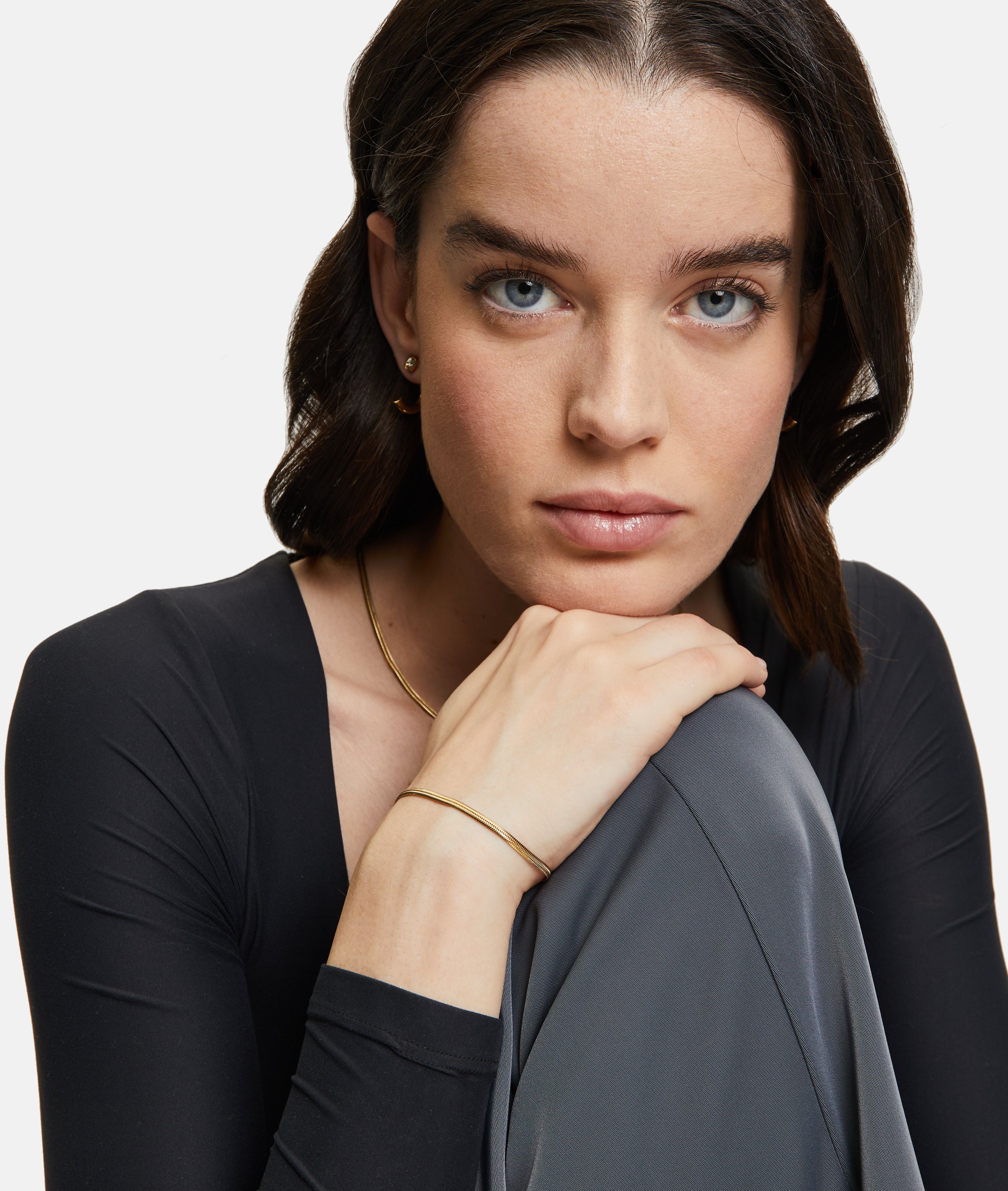 Frau mit blauen Augen und dunklem Haar stützt ihr Kinn auf die Hand. Sie trägt Goldschmuck - eine Schlangenkette und ein elegantes Armband - und ein schwarzes Tamaris Set - The Layer Top.