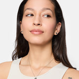 Frau mit langen braunen Haaren, trägt ein weißes Tanktop, Ohrringe, Damen Schmuck, eine Tamaris Halskette - Modern Layering von Tamaris und ein m-Tattoo auf ihrer Schulter.
