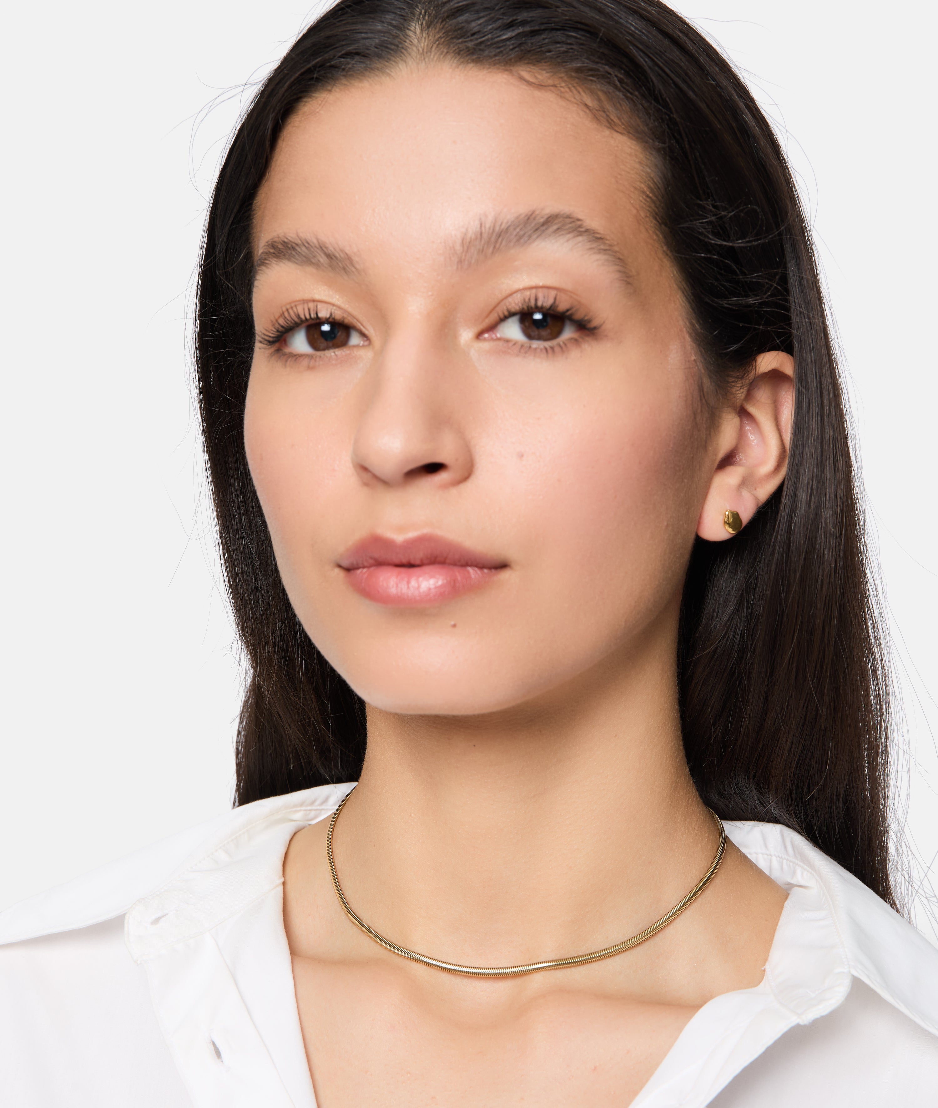 Eine Frau mit langen dunklen Haaren trägt ein weißes Hemd, goldene Ohrringe und die Tamaris Halskette - Modern Layering von Tamaris und schaut in die Kamera.