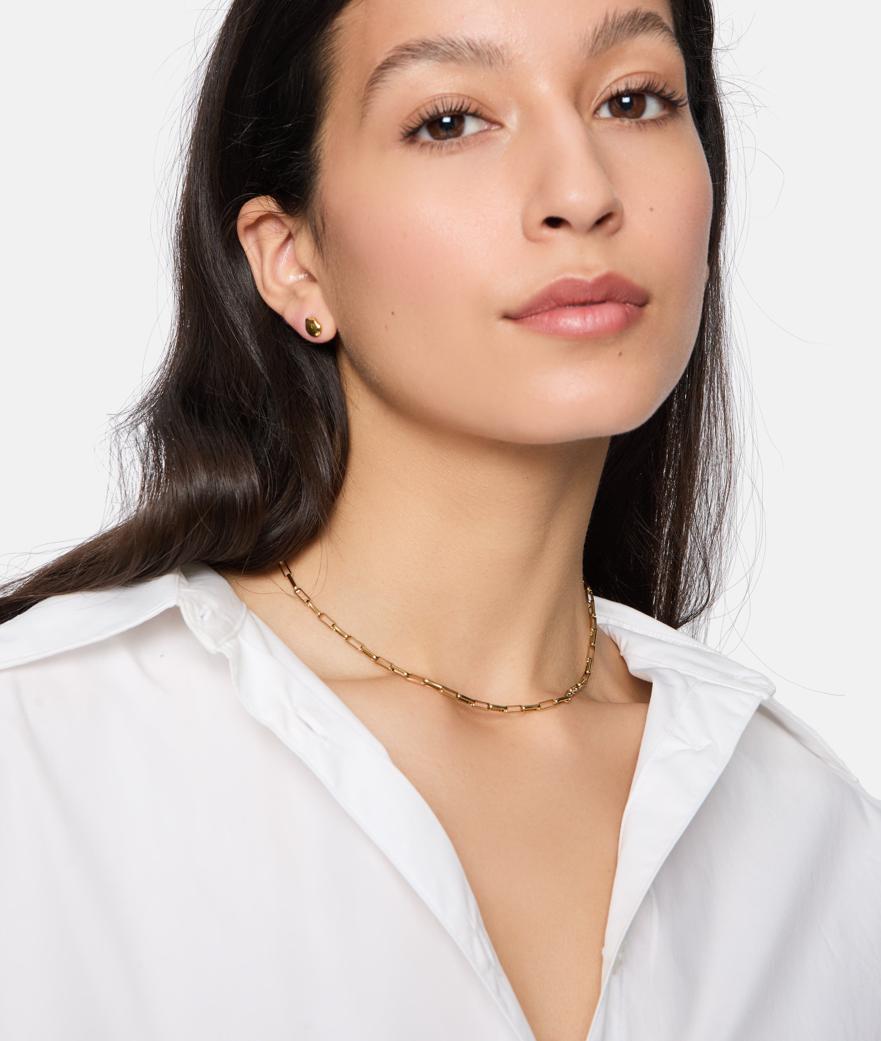 Frau in weißem Hemd mit Tamaris Halskette - Modern Layering und kleinen goldenen Ohrringen. Ihr glattes dunkles Haar vervollständigt einen eleganten Look mit Damen Schmuck und dezent gestufter Schlangenkette.