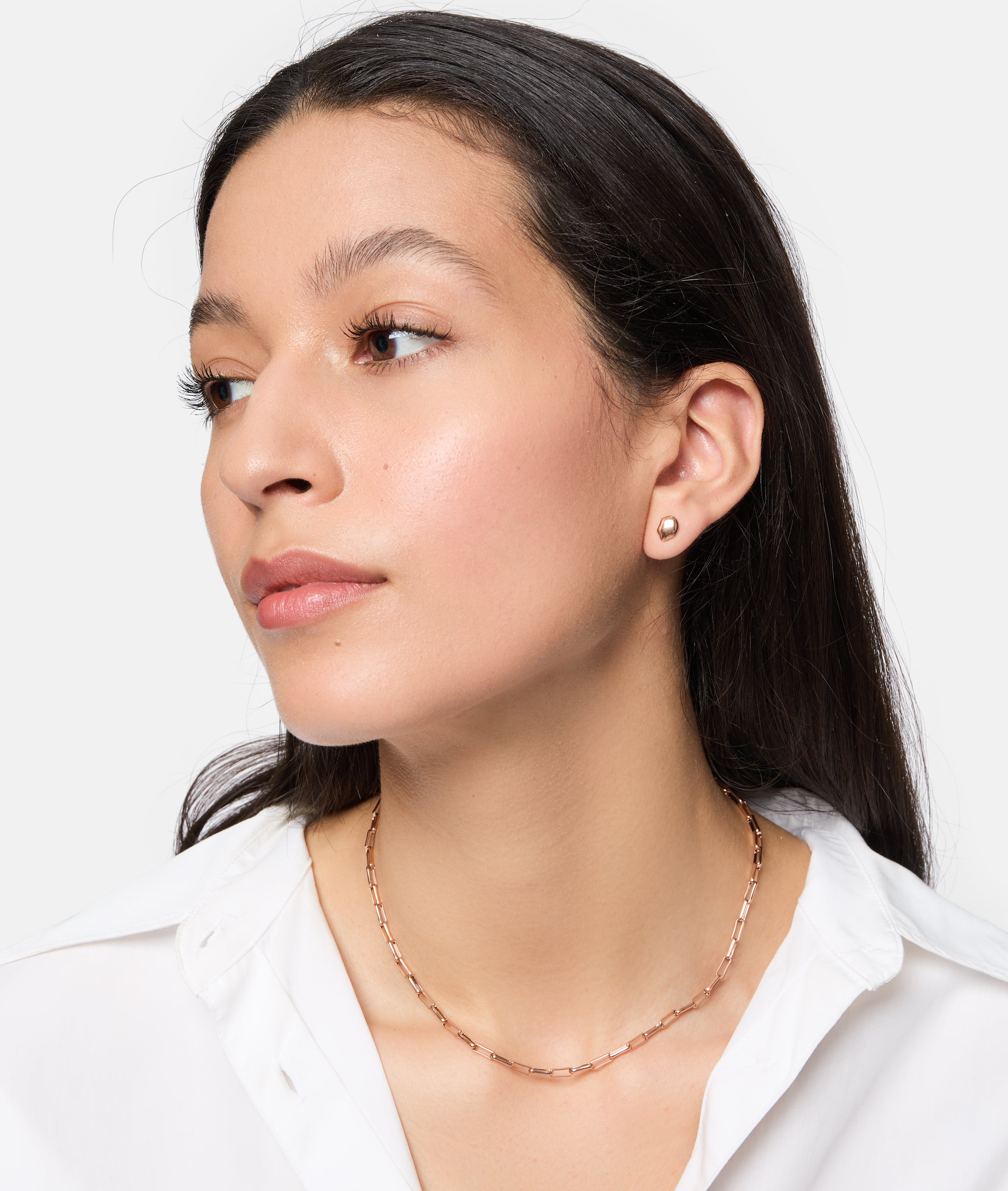 Frau mit langen dunklen Haaren trägt ein weißes Hemd und die Tamaris Halskette - Modern Layering von Tamaris, gepaart mit kleinen Ohrsteckern, mit Blick zur Seite.