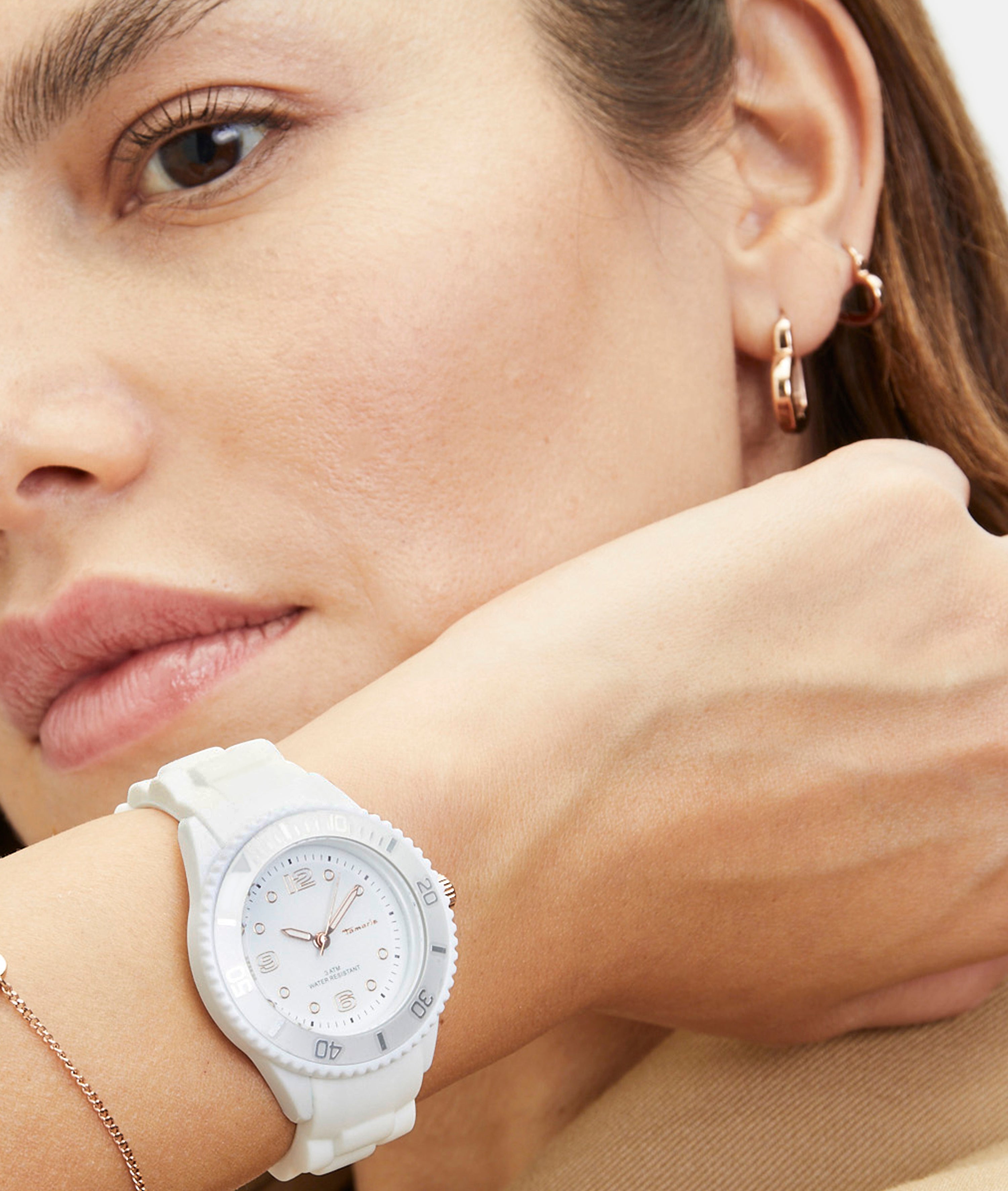 Eine Frau, die eine Tamaris Armbanduhr - The White Silicone und goldene Ohrringe trägt, stützt ihre Hand auf ihr Gesicht.