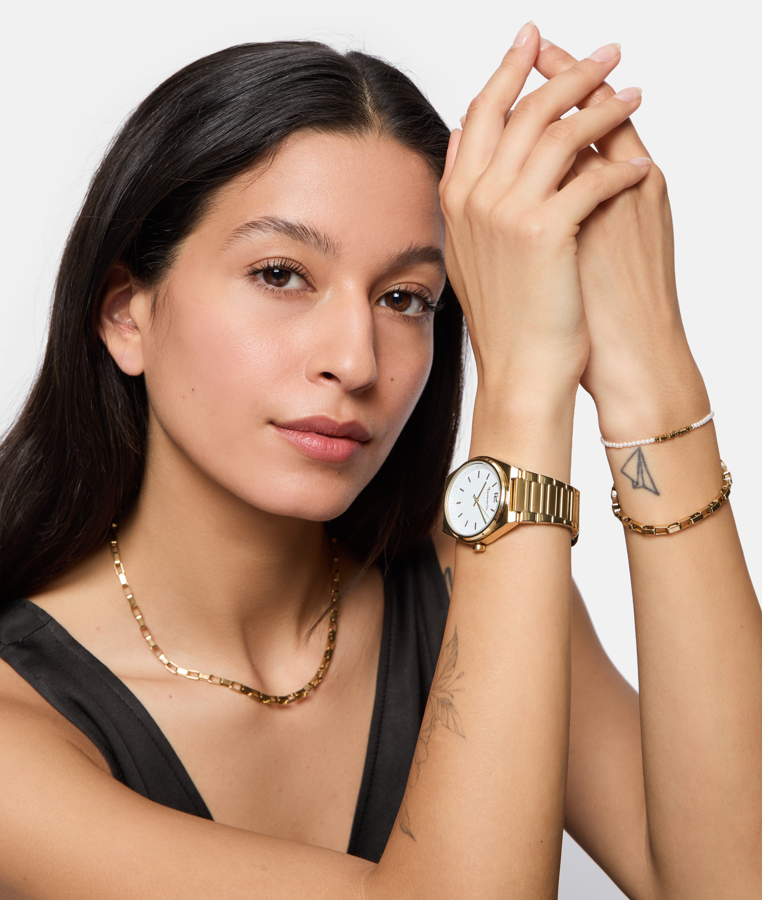 Eine Frau mit langen dunklen Haaren posiert und trägt eine goldene Uhr, eine Kettenhalskette und das Tamaris Armband - Modern Pearl Fusion von Tamaris; ihre Tätowierungen sind deutlich sichtbar.