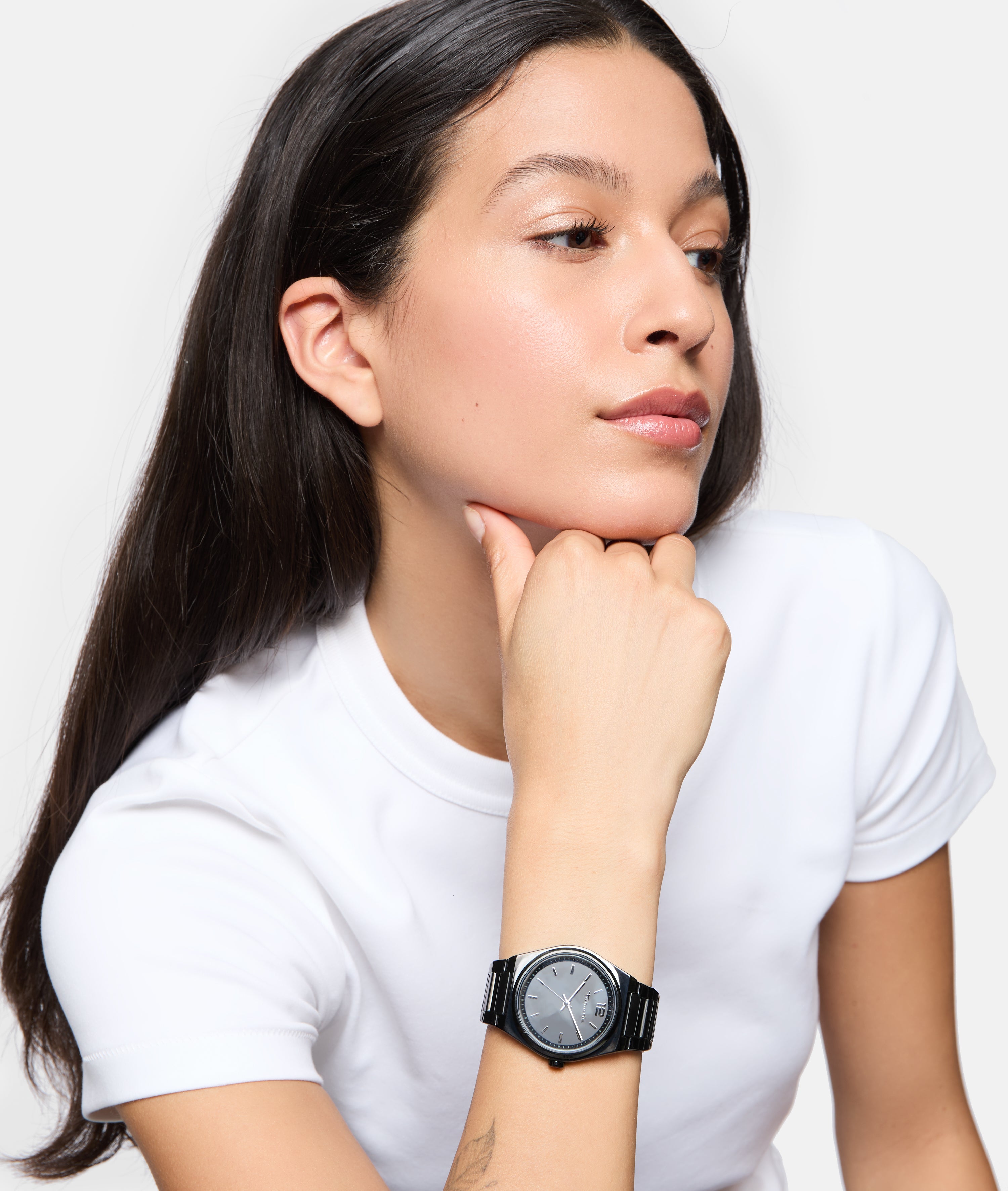 Eine Frau im weißen Hemd stützt gedankenverloren ihr Kinn auf die Hand und trägt die Tamaris Armbanduhr - The Round Basic Essential, die zeitlose Eleganz ausstrahlt.