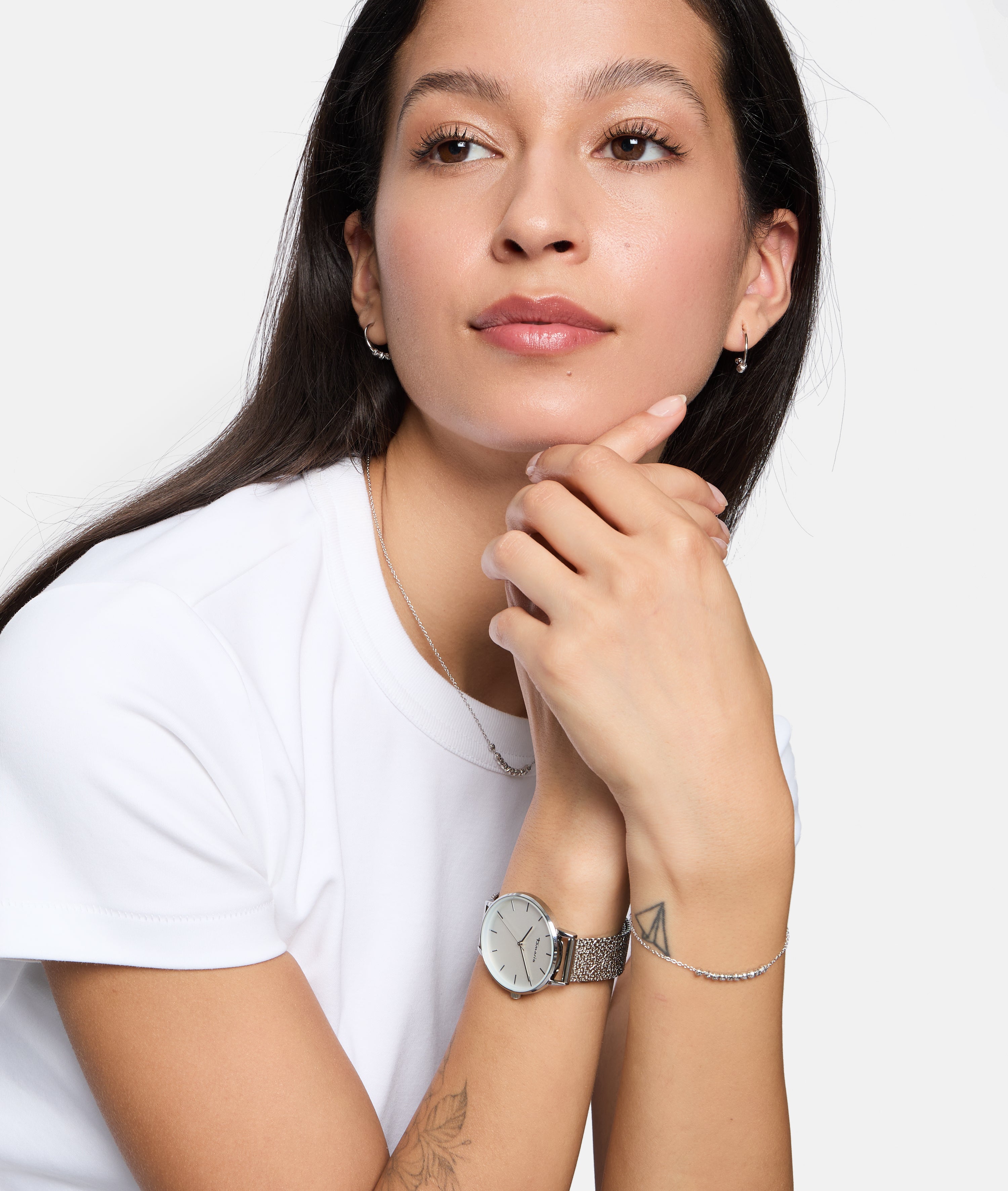Eine Frau in einem weißen Hemd posiert mit verschränkten Händen und hebt das Tamaris Armband - The Glimmer und eine silberne Uhr als Teil ihres Schmucks vor einem weißen Hintergrund hervor.