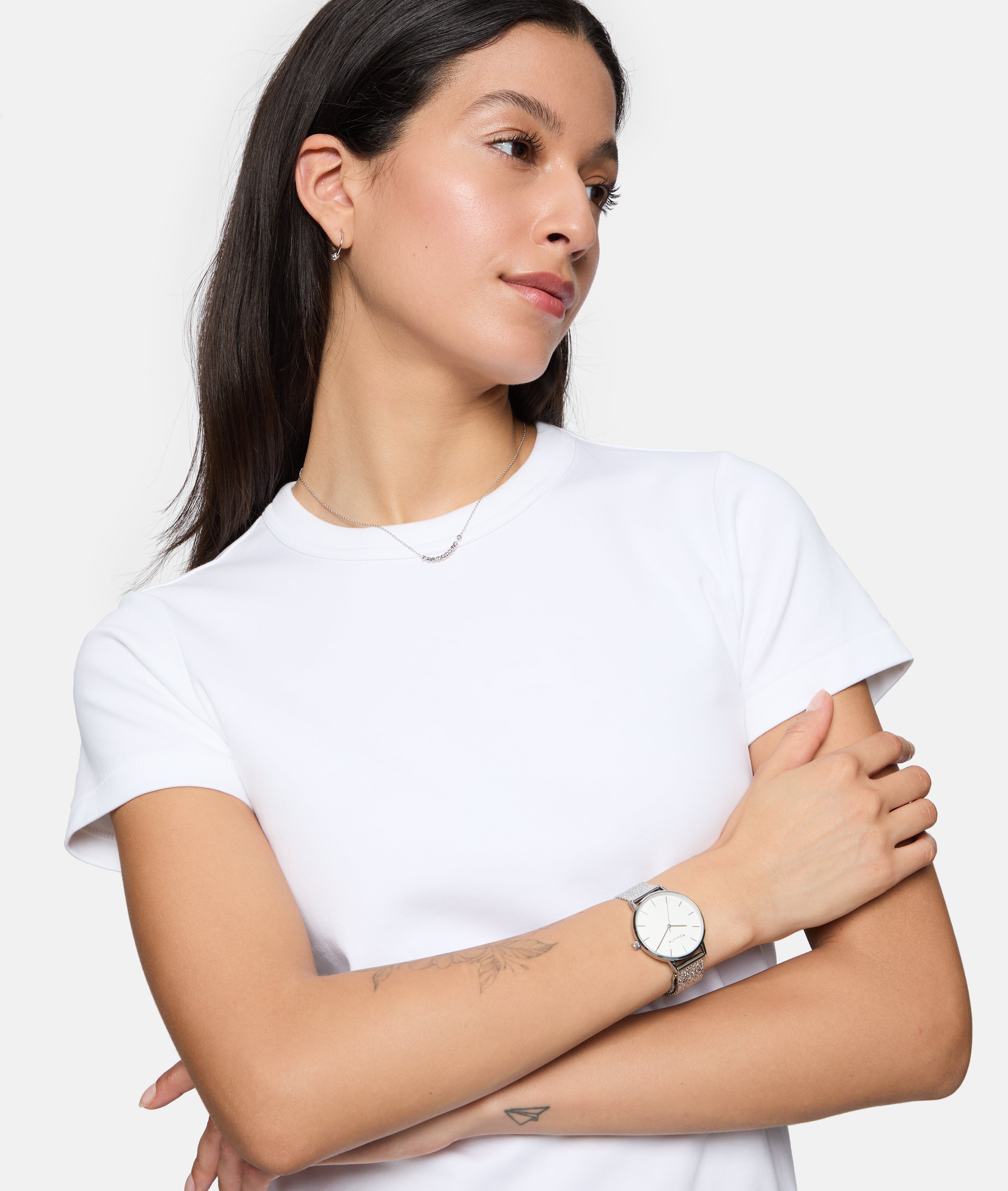 Frau mit dunklen Haaren in einem weißen T-Shirt, Arme verschränkt, trägt die Tamaris Armbanduhr - Sparkle Up von Tamaris mit einem Netzband, schaut zur Seite.