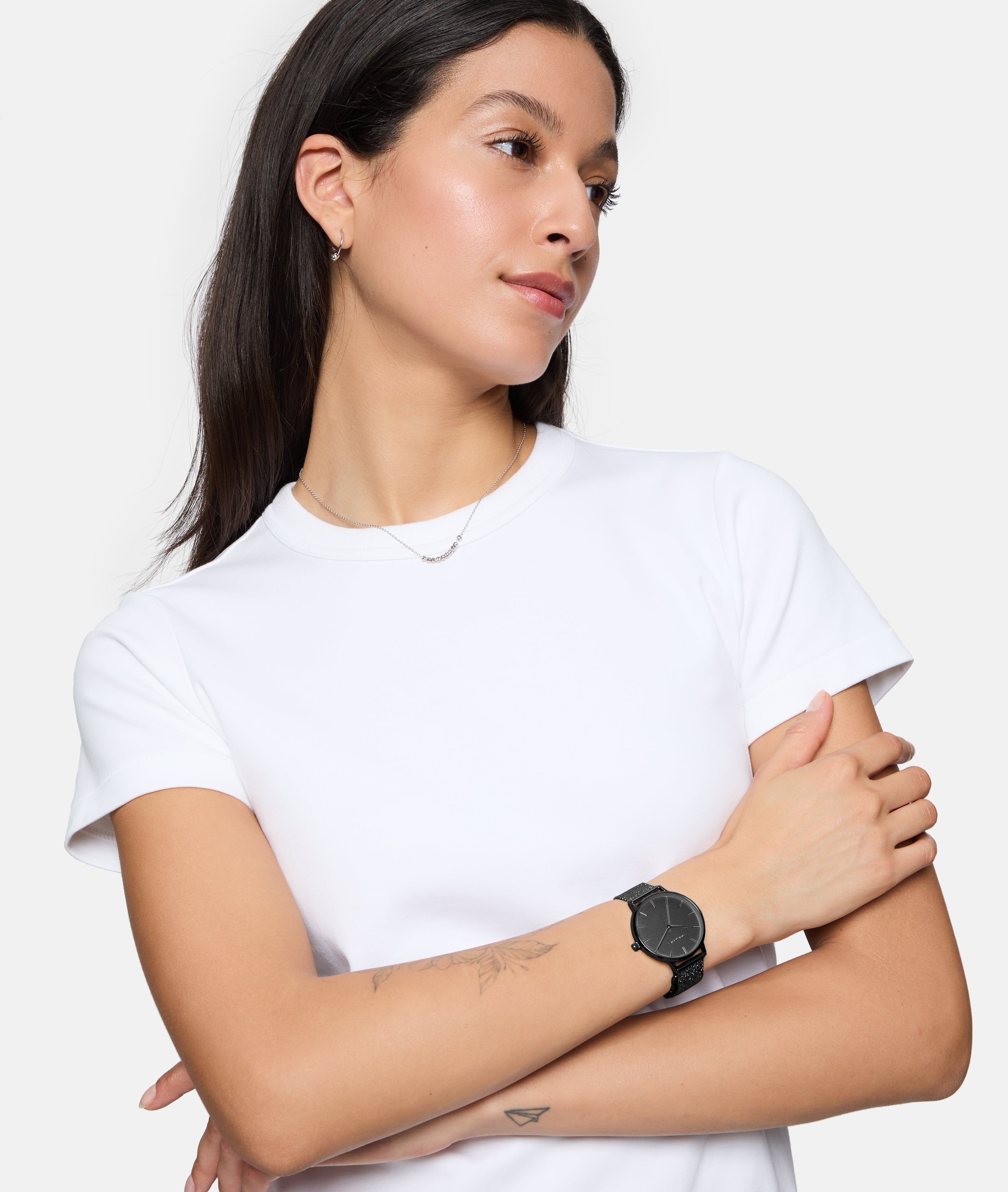 Eine Frau in einem weißen T-Shirt trägt die Tamaris Armbanduhr - Sparkle Up mit elegantem Design und schlankem Meshband am Handgelenk und schaut mit verschränkten Armen zur Seite.