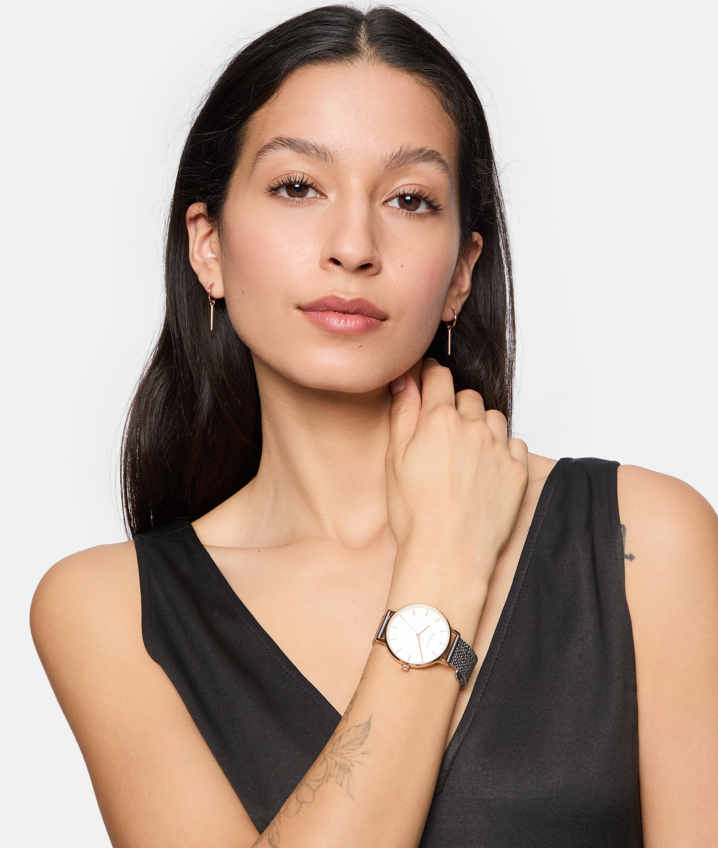 Eine Frau mit langen dunklen Haaren, einem schwarzen ärmellosen Oberteil und kleinen Ohrringen posiert selbstbewusst und trägt die Tamaris Armbanduhr - Sparkle Up by Tamaris mit einem stilvollen Netzband.