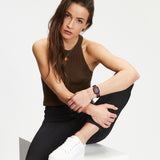 Eine Frau in einem braunen Tank-Top und einer schwarzen Hose sitzt auf einem weißen Block, ein Knie hoch, und schaut in die Kamera. Sie präsentiert die stylische Tamaris Armbanduhr - The Colorful Square von Tamaris.