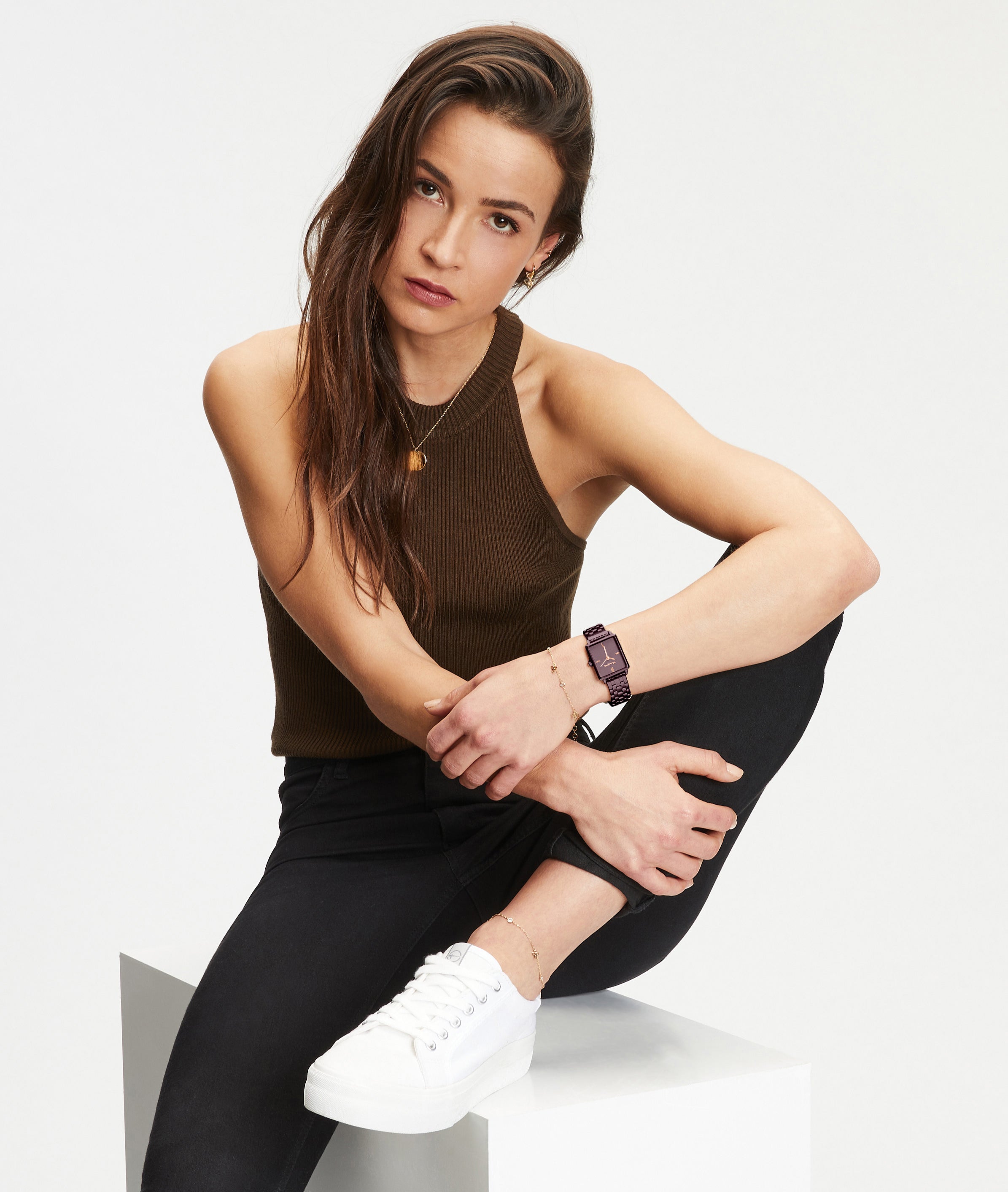Eine Frau in einem braunen Tank-Top und einer schwarzen Hose sitzt auf einem weißen Block, ein Knie hoch, und schaut in die Kamera. Sie präsentiert die stylische Tamaris Armbanduhr - The Colorful Square von Tamaris.