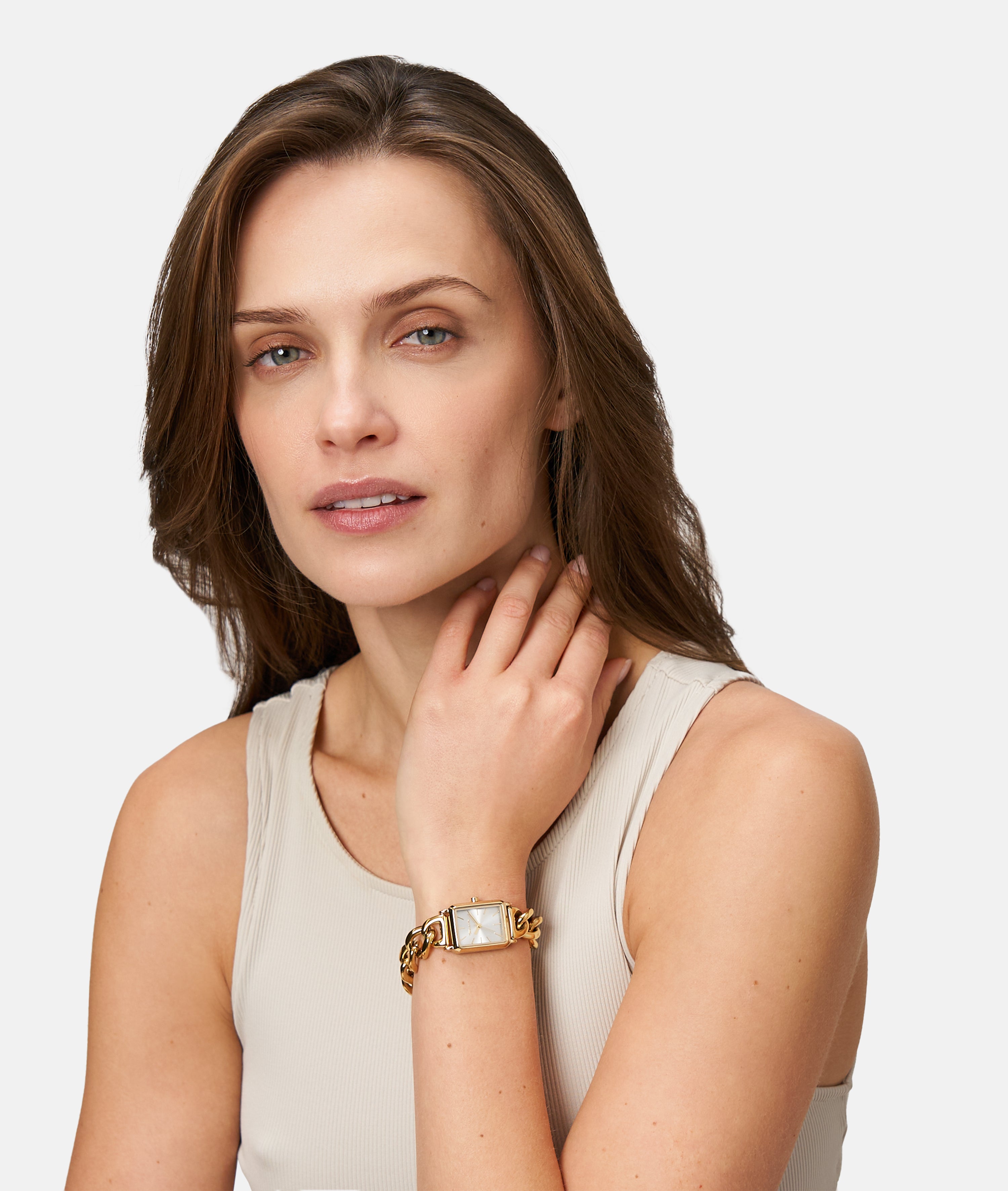 Frau mit hellbraunem Haar, die ein beiges Tank-Top und eine Tamaris Armbanduhr - Chain Watch von Tamaris trägt, berührt ihren Hals und schaut nachdenklich in die Kamera.
