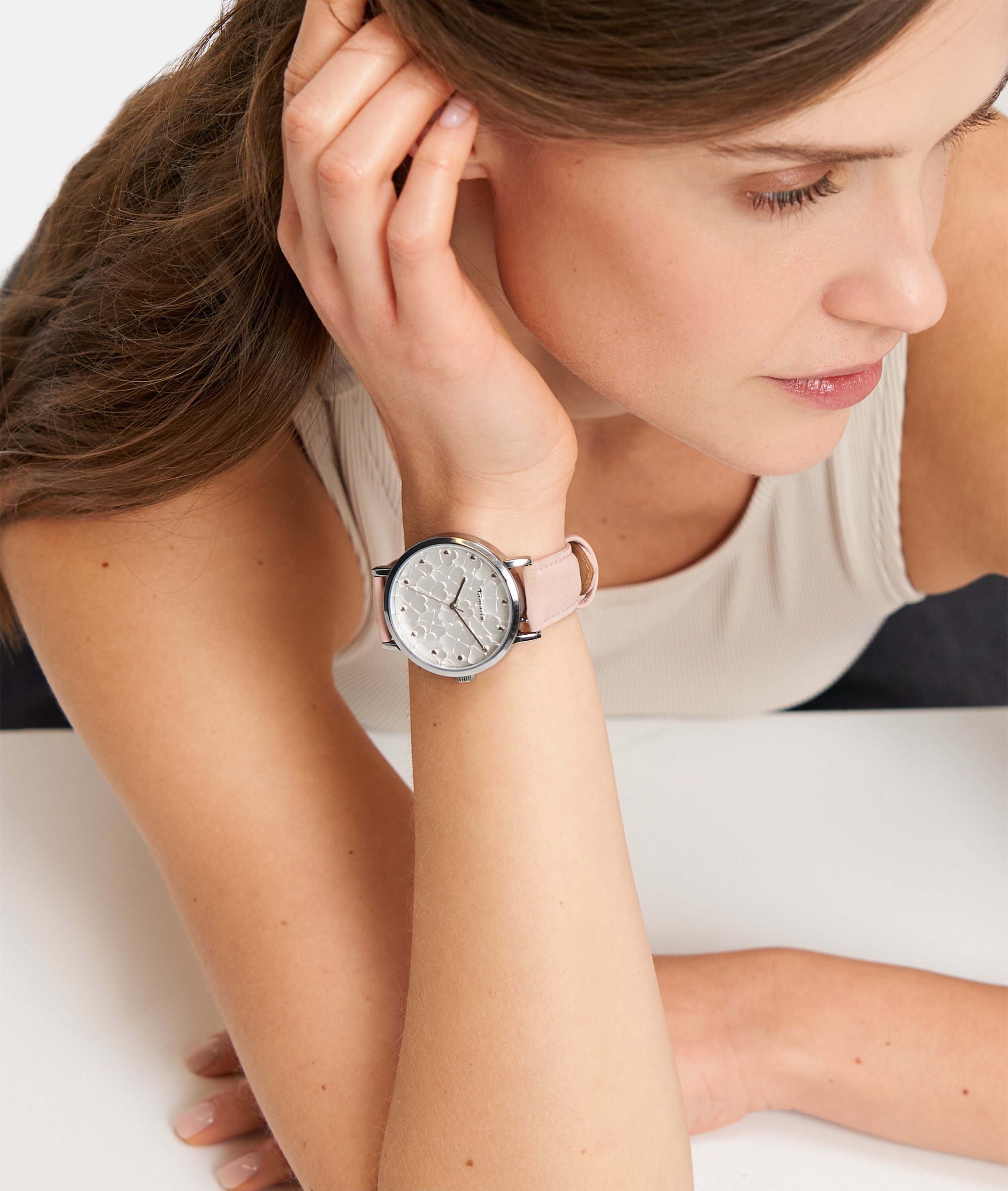 Eine Frau in einem ärmellosen Top stützt ihren Kopf auf die Hand und trägt die Tamaris Armbanduhr - Clover mit floralem Zifferblatt und stilvollem rosa Lederband von Tamaris am Handgelenk.