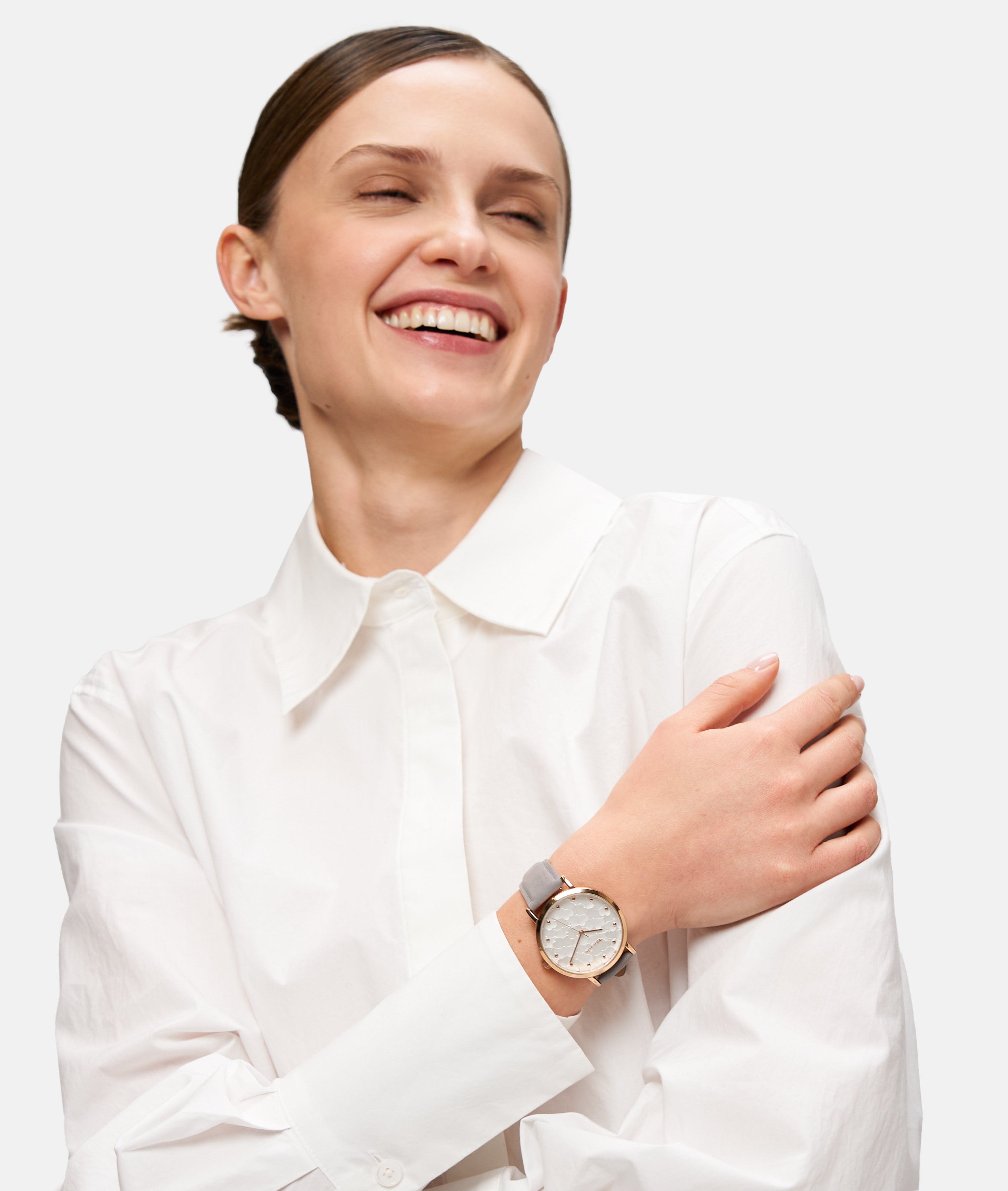 Lächelnde Frau in weißem Hemd mit verschränkten Armen, trägt die Tamaris Armbanduhr - Clover von Tamaris mit hellem Band und floralem Zifferblatt.