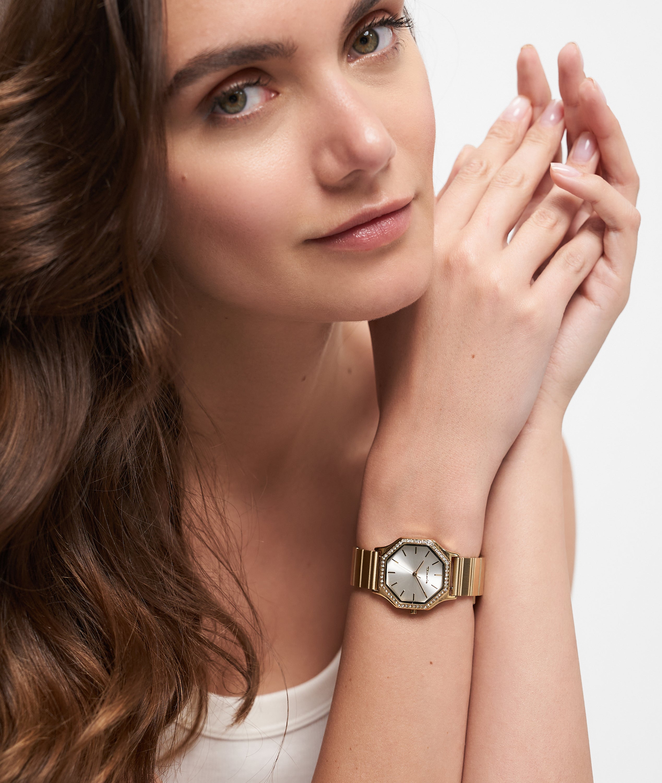 Frau mit langen braunen Haaren trägt eine Tamaris Sparkling Octavia Armbanduhr mit schimmernden Zirkoniasteinen und goldenem Edelstahlband, die Hände sanft in ihrem weißen Tanktop verschränkt.