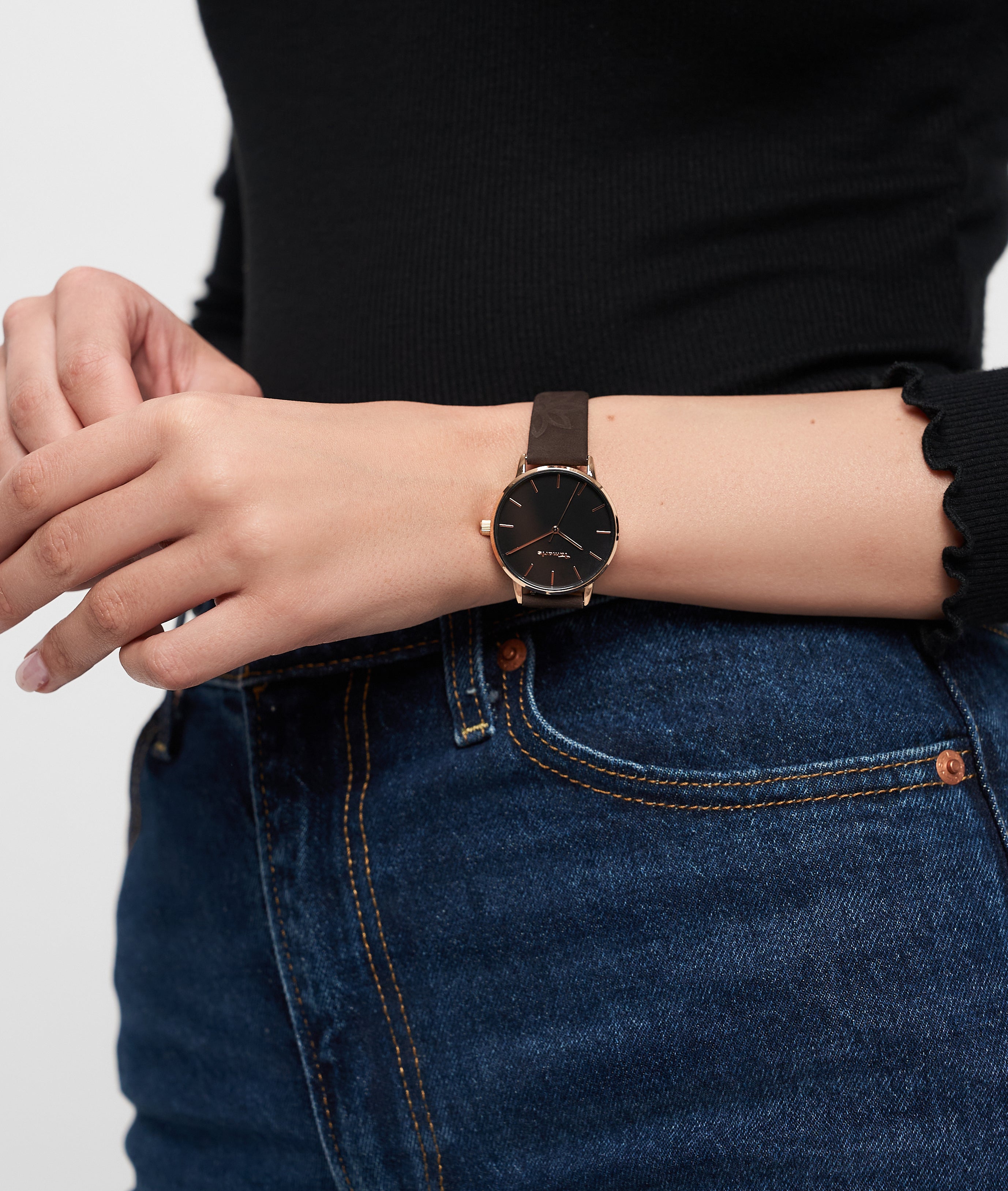 Eine Person, die eine Tamaris Armbanduhr - Stamped Flowers, ein schwarzes langärmeliges Oberteil und eine blaue Jeans trägt.