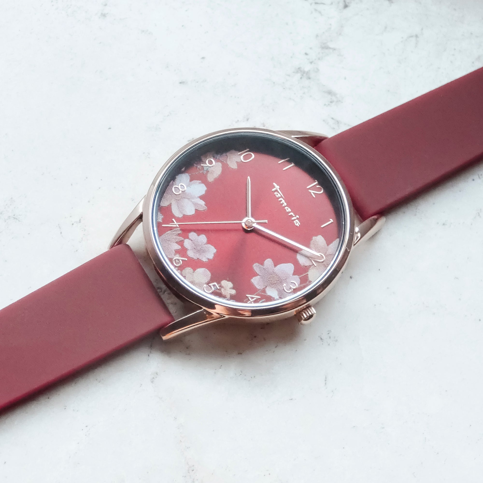 Eine Tamaris Armbanduhr - Sporty Flowers von Tamaris mit kastanienbraunem Silikonarmband und rotem Blumenmotiv wird auf einer hellen Marmorfläche präsentiert.