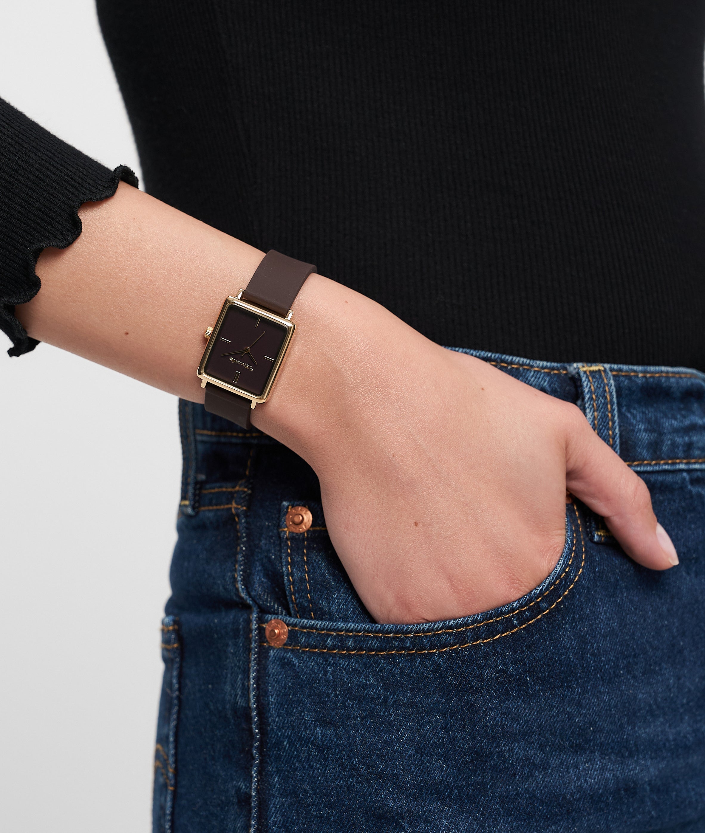 Eine Person trägt ein schwarzes langärmeliges Oberteil, blaue Jeans und eine sportliche Tamaris Armbanduhr - The Silicon Square: eine quadratische schwarze Uhr mit goldenem Rahmen von Tamaris.