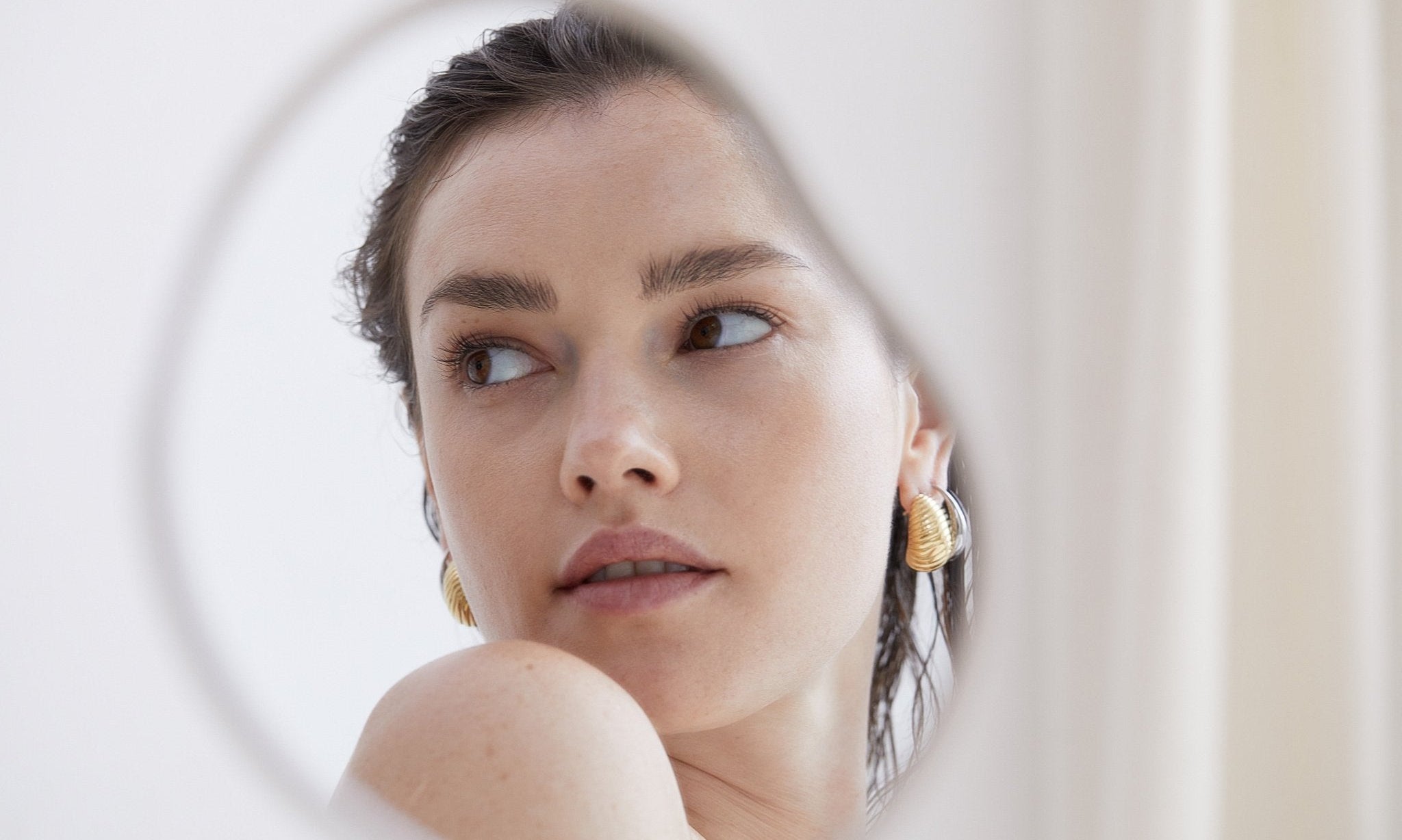 Frau mit goldenen Echtzeit-Creolen im Spiegelbild – moderner Schmuck trifft auf zarte Eleganz und minimalistisches Design.