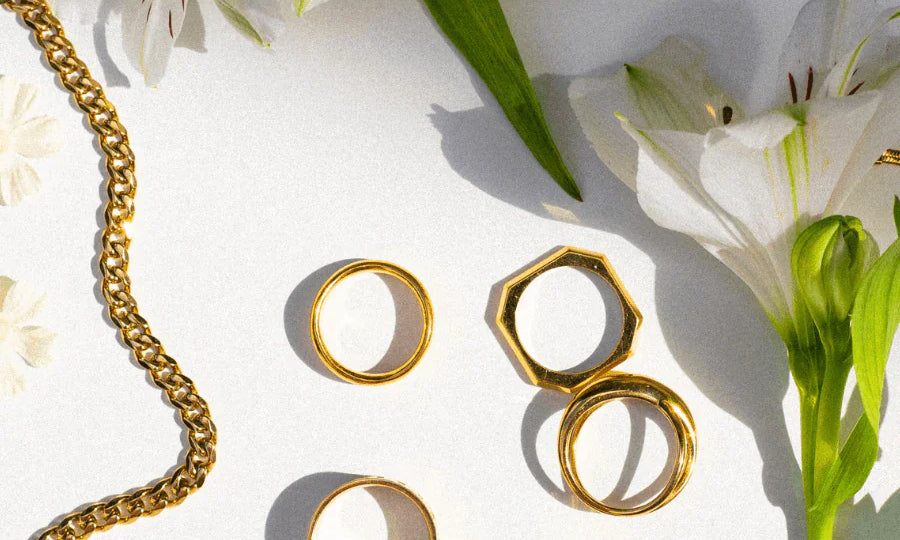 Stimmungsvolles Flatlay mit goldfarbenem Schmuck von Tamaris und Liebeskind Berlin, dekorativ arrangiert zwischen weißen Blumen im Sonnenlicht.