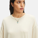 Frau in einem lockeren cremefarbenen Hemd, die geschichtete silberne Liebeskind Berlin Halsketten trägt, darunter "The Colour Epoxy" mit einem T-Bar-Verschluss und achteckigem Anhänger, mit Blick zur Seite.