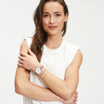 Eine Frau mit langen braunen Haaren in einem weißen ärmellosen Oberteil, die eine Armbanduhr trägt und in die Kamera lächelt, präsentiert elegante Tamaris Ohrhänger - Flower Ohrringe.
