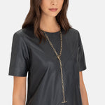 Eine Frau in einem schwarzen kurzärmeligen Lederkleid trägt die Liebeskind Berlin Halskette - The long T-bar necklace vor einem schlichten Hintergrund.