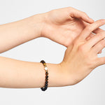 Eine Hand, die das LIEBESKIND BERLIN Beads - Onyx meets Tigers Eye Armband von Liebeskind Berlin mit eleganten schwarzen und goldenen Perlen trägt, ruht vor einem schlichten, hellen Hintergrund, um dieses stilvolle Schmuck Accessoire hervorzuheben.
