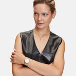 Eine Frau in einem schwarzen ärmellosen Ledertop verschränkt die Arme und trägt mehrere goldene Halsketten, darunter die Liebeskind Berlin Halskette - The Sleek, sowie eine goldene Uhr, um einen schicken und modernen Look zu kreieren.