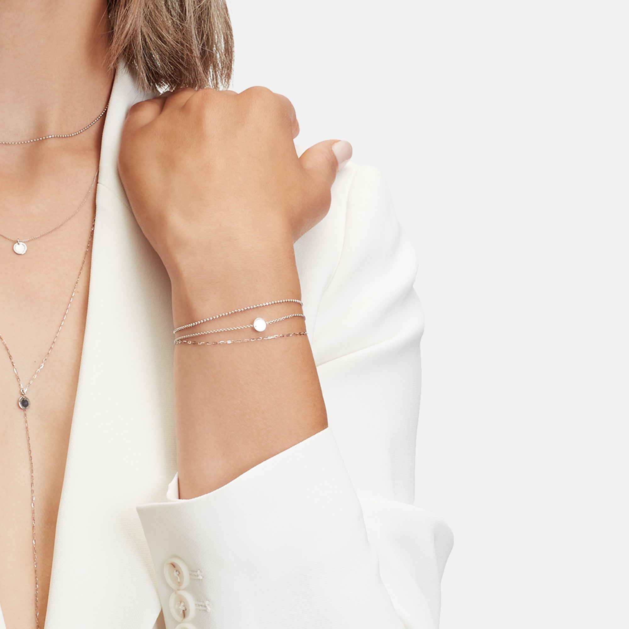 Eine Frau in einem weißen Blazer trägt das Gold Collection Armband von Liebeskind Berlin, das mit silbernen Halsketten und Armbändern mit kleinen runden Anhängern kombiniert ist.