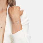 Eine Frau in einem weißen Blazer trägt zarten Silberschmuck und das LIEBESKIND BERLIN Armband - Gold Collection von Liebeskind Berlin. Es besteht aus mehrlagigen Halsketten und schmalen Armbändern mit kleinen runden Anhängern vor einem neutralen Hintergrund.
