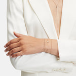 Eine Person im weißen Anzug mit verschränkten Armen präsentiert zarten Schmuck aus 18 Karat Roségold, darunter eine lange Halskette und zwei minimalistische LIEBESKIND BERLIN Armbänder aus der Liebeskind Berlin Gold Collection, die jeweils kleine runde Akzente aufweisen.
