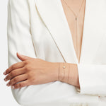 Eine Person in einem weißen Blazer mit tiefem Dekolleté präsentiert mehrlagigen Goldschmuck aus der Liebeskind Berlin Gold Collection: eine Halskette aus 18 Karat Roségold und zwei zarte LIEBESKIND BERLIN Armbänder, darunter eines mit einem runden Anhänger, mit verschränkten Armen.