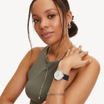 Frau in einem ärmellosen grünen Top trägt eine silberne Tamaris Armbanduhr - The Classic Canvas und eine Halskette sowie ein Armband und posiert vor einem weißen Hintergrund - ein echter Basic Must-Have-Look.